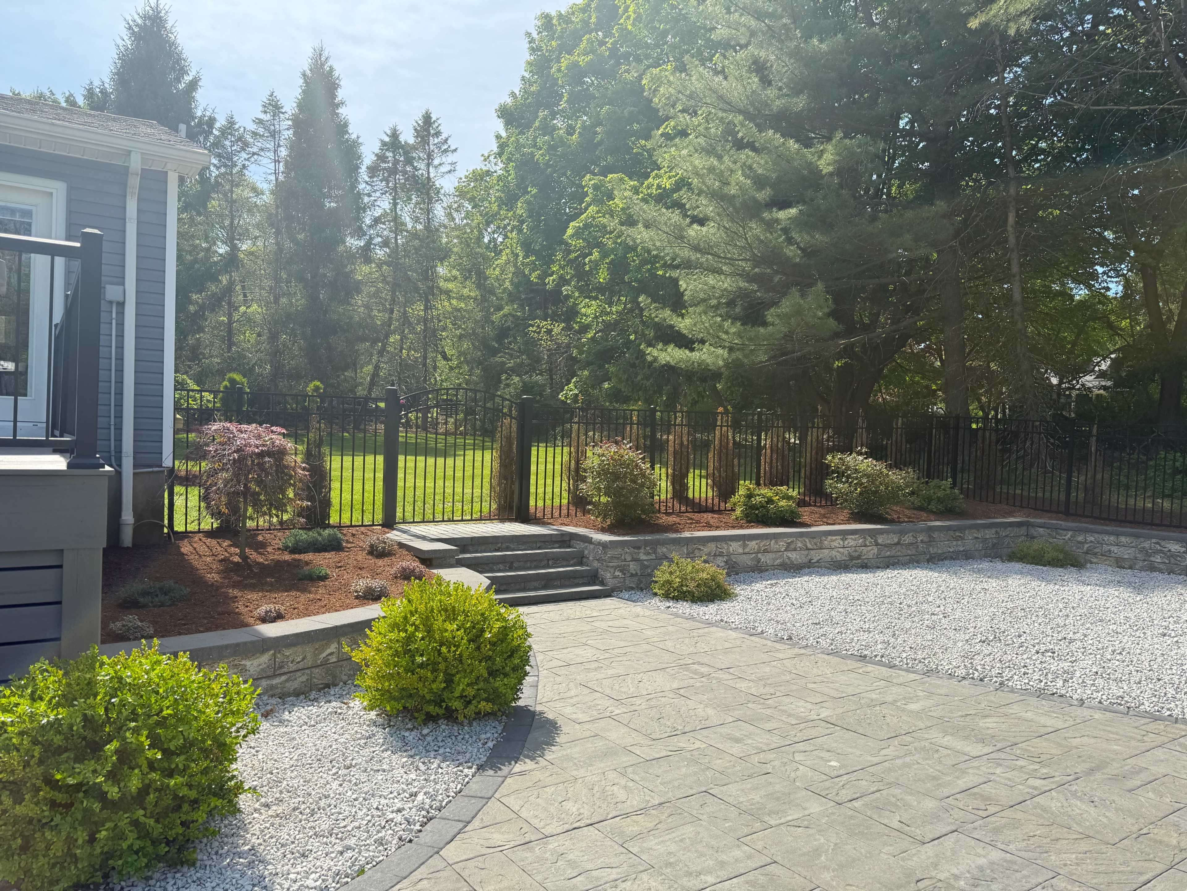 The image shows a landscaped yard with a stone pathway, gravel surface, and a fenced area featuring green grass and trees in the background.