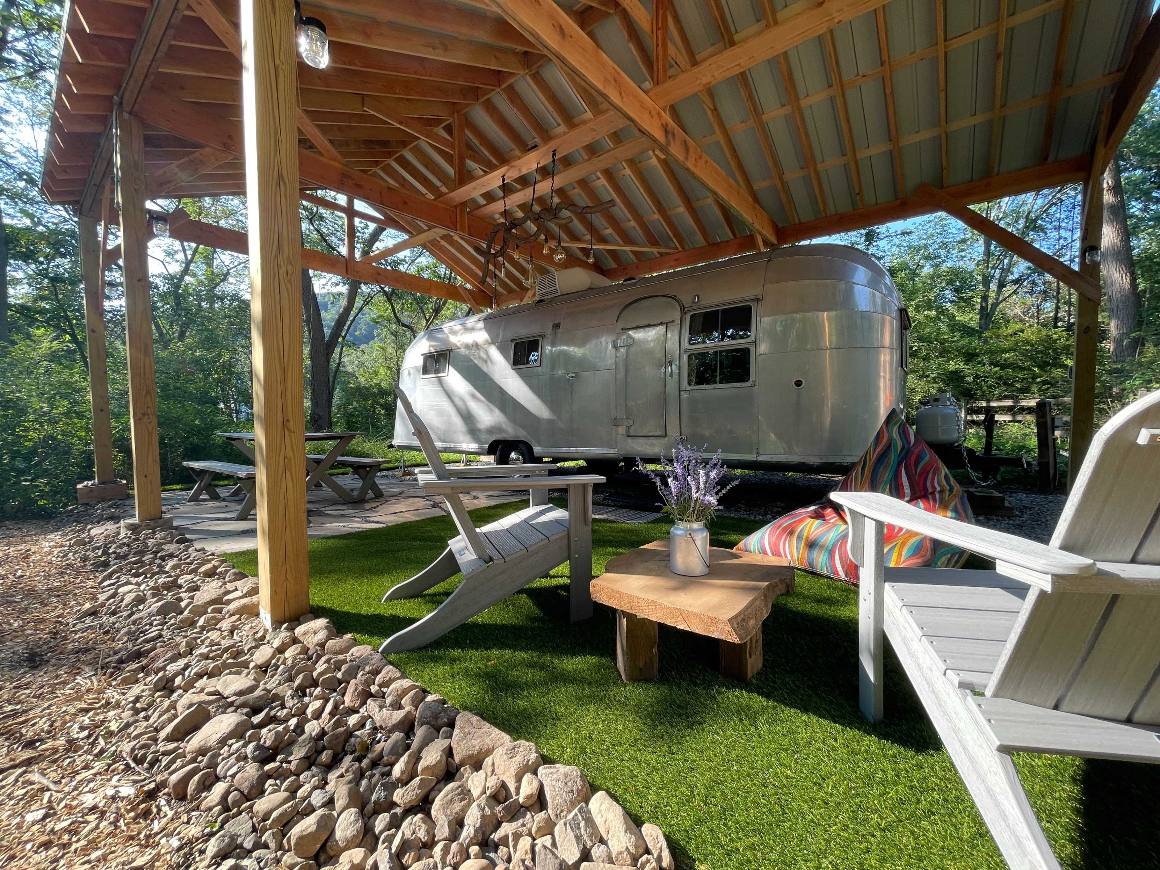 A vintage Airstream trailer is parked under a wooden shelter surrounded by a grassy area and decorative rocks.
