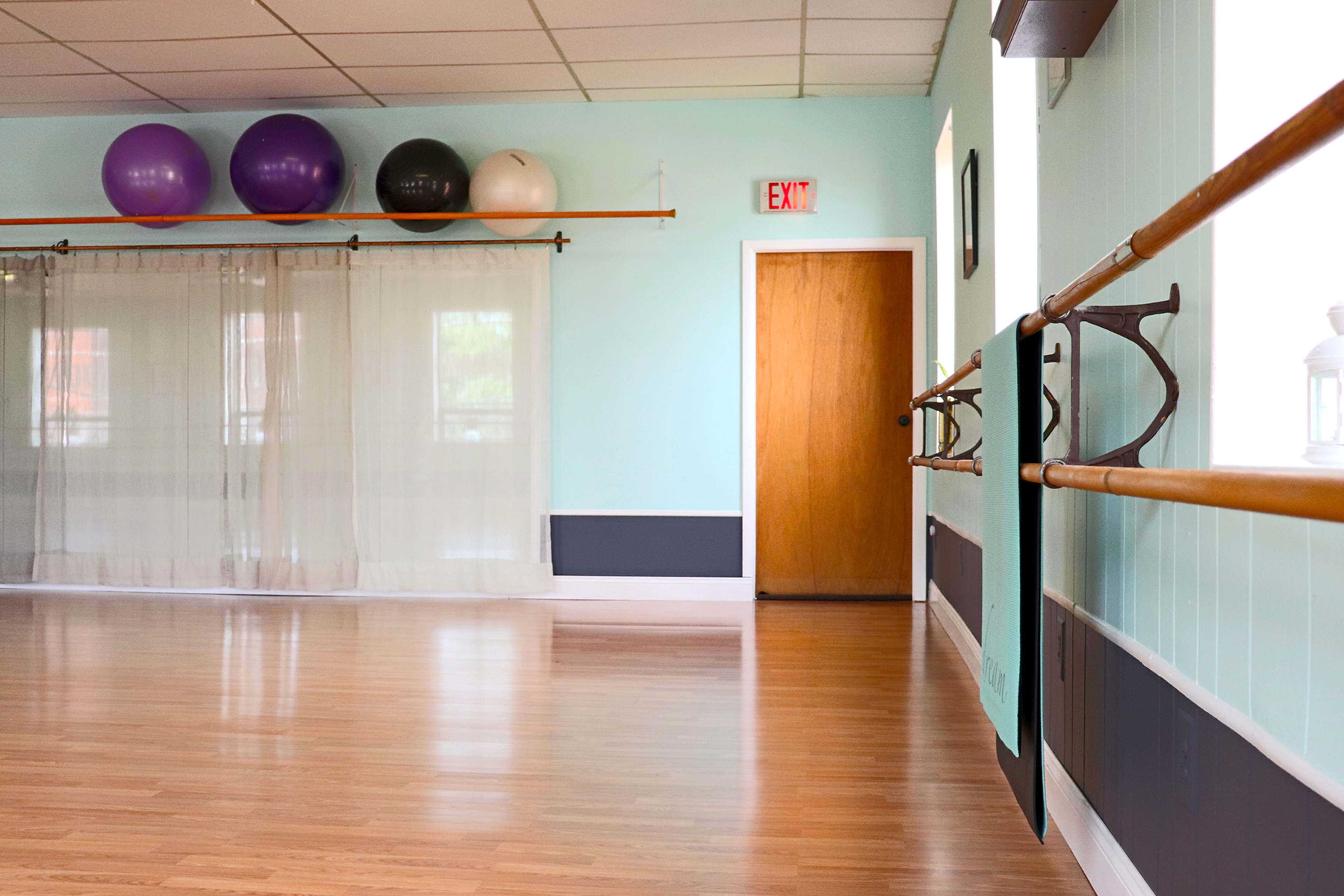 The image shows a light blue dance studio with exercise balls arranged on a shelf, wooden flooring, and a door leading to another room.