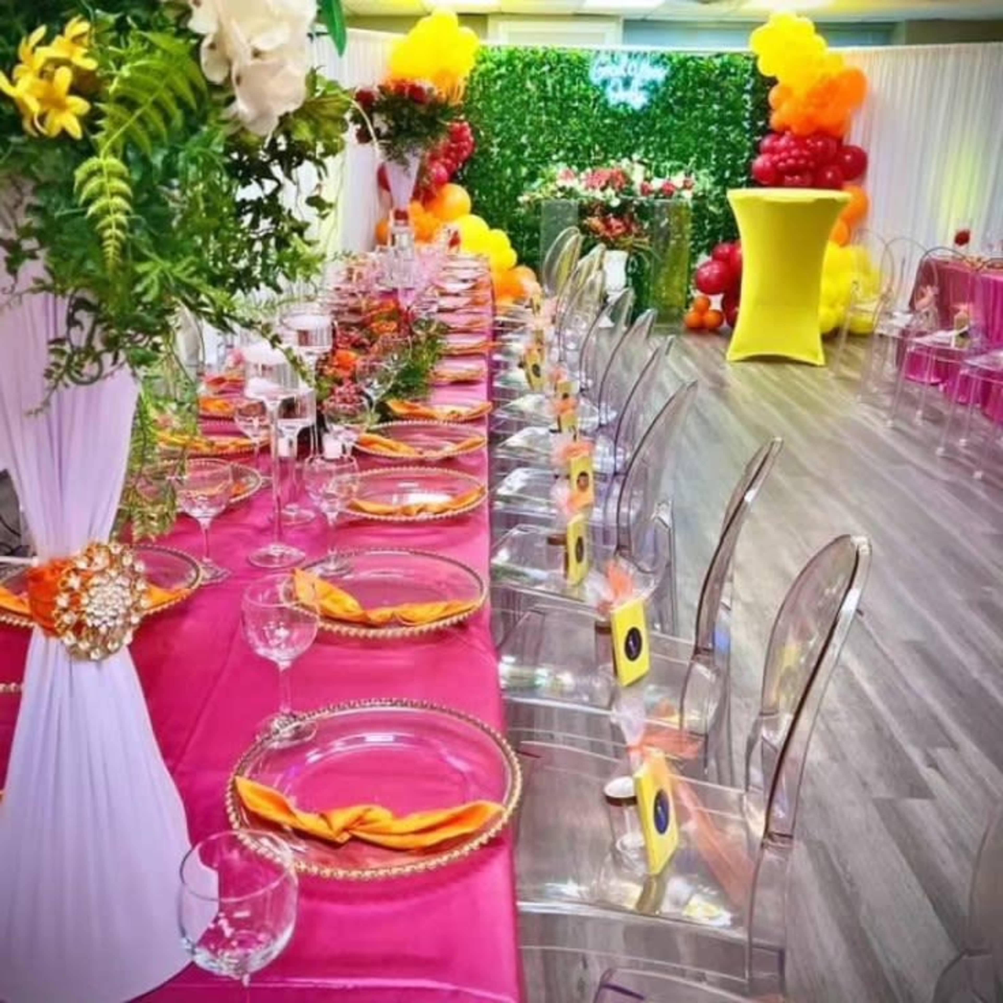A long dining table is set with colorful tablecloths, clear chairs, and decorative centerpieces in a vibrant event space.