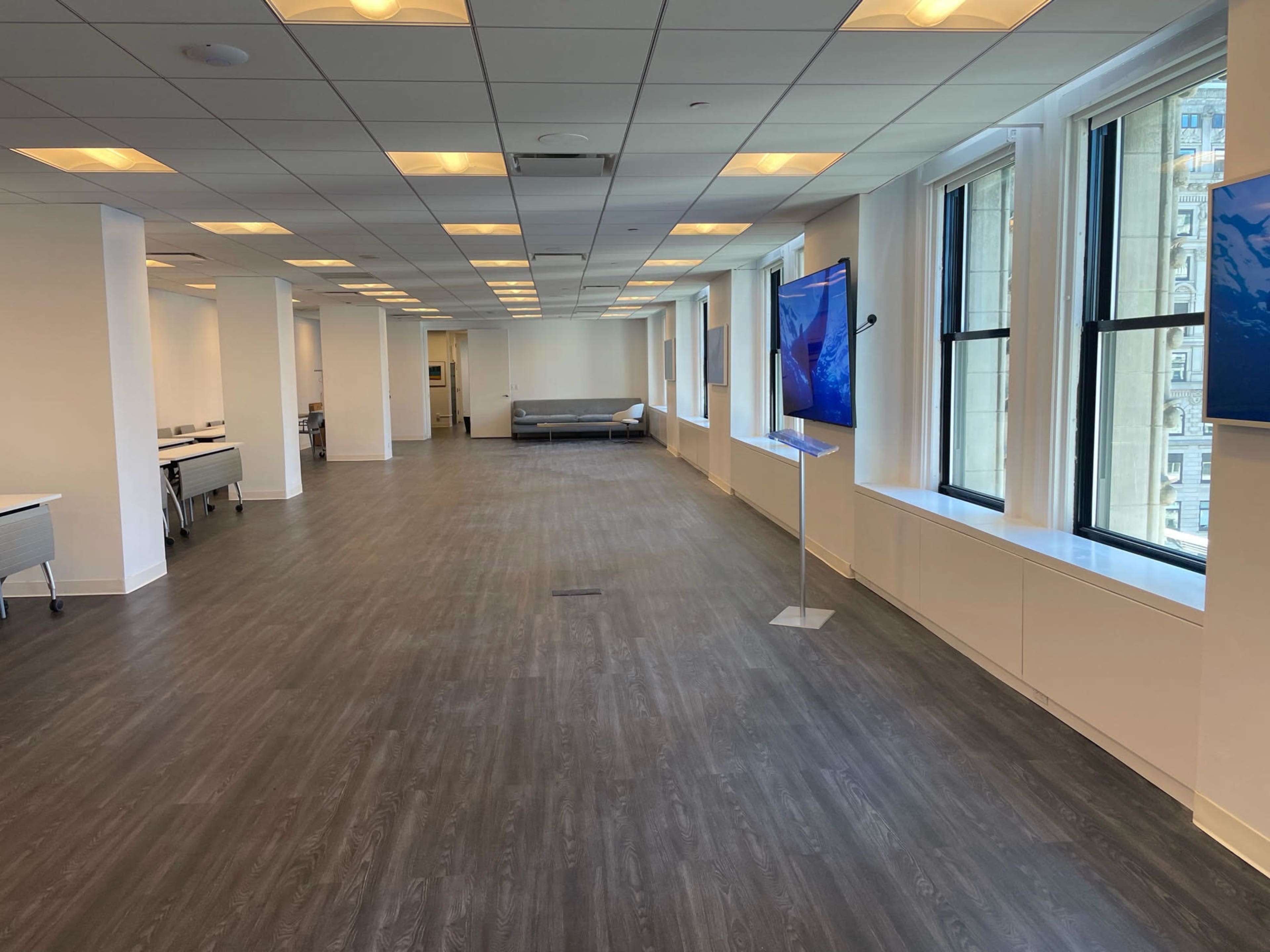 A spacious, brightly lit office area with large windows and a polished wooden floor, featuring a seating area and two mounted screens.