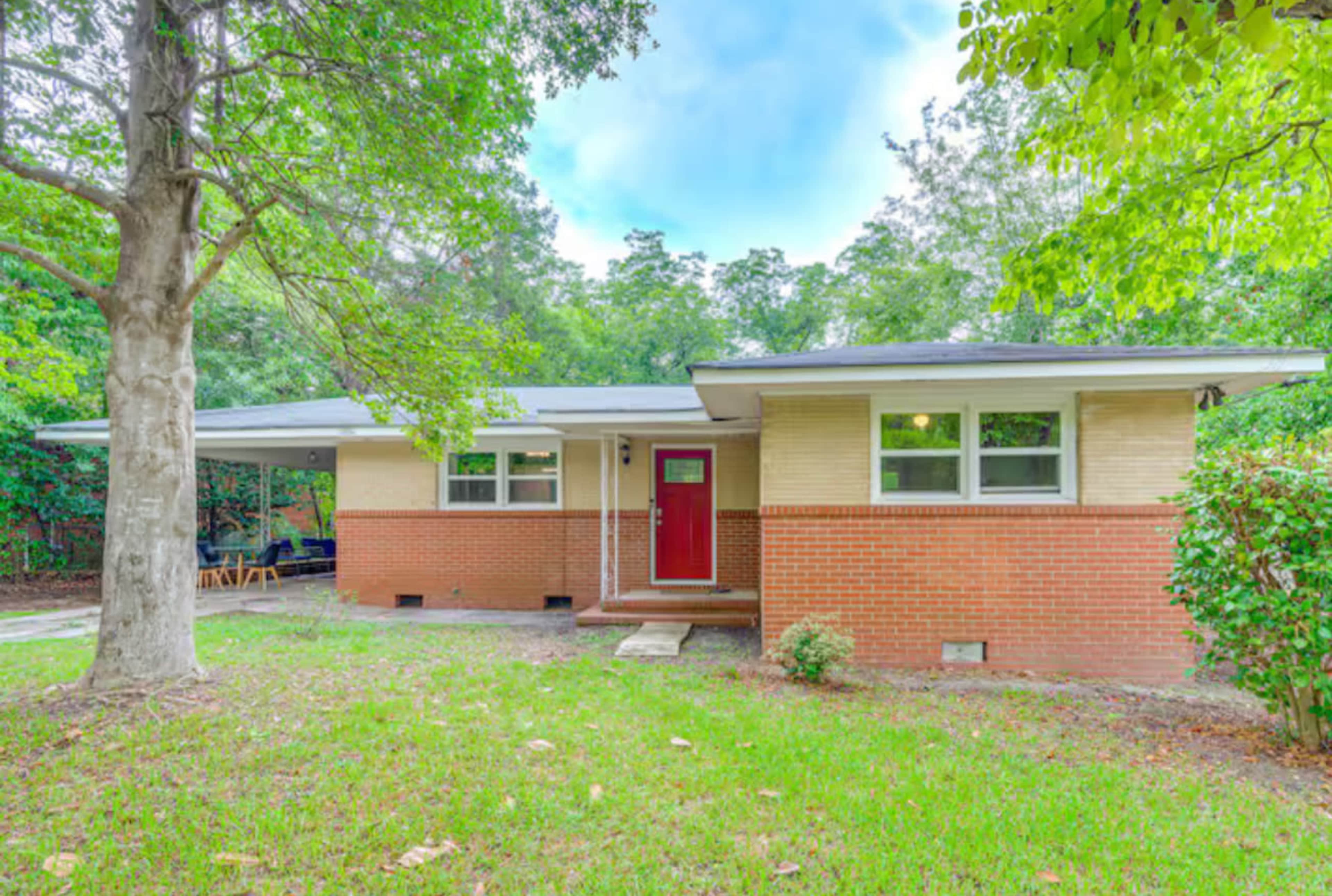 A single-story brick house with a red front door sits in a lush, green yard surrounded by trees.