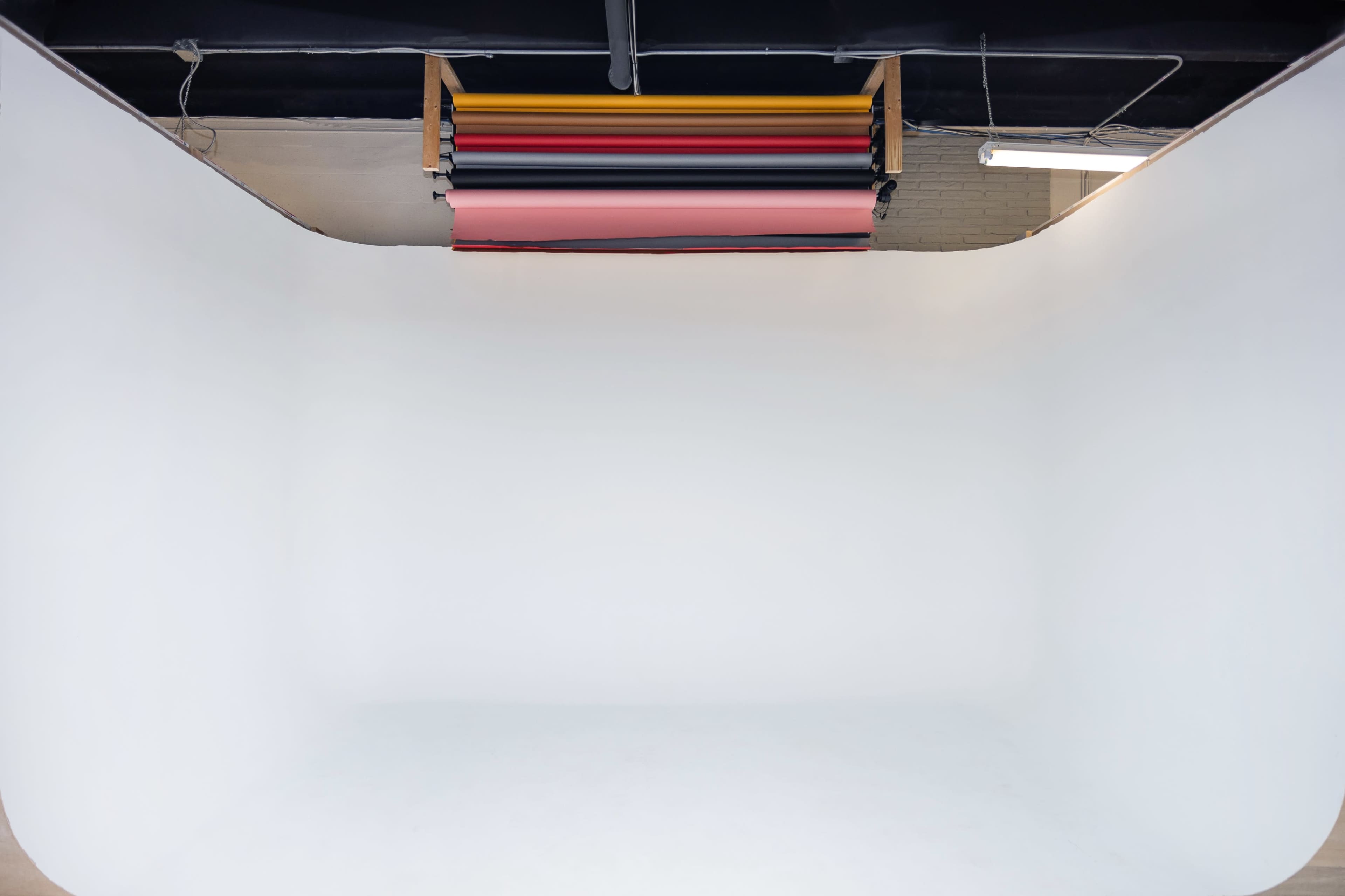 The image shows an empty photography studio with a white backdrop and colored paper rolls hanging from the ceiling.