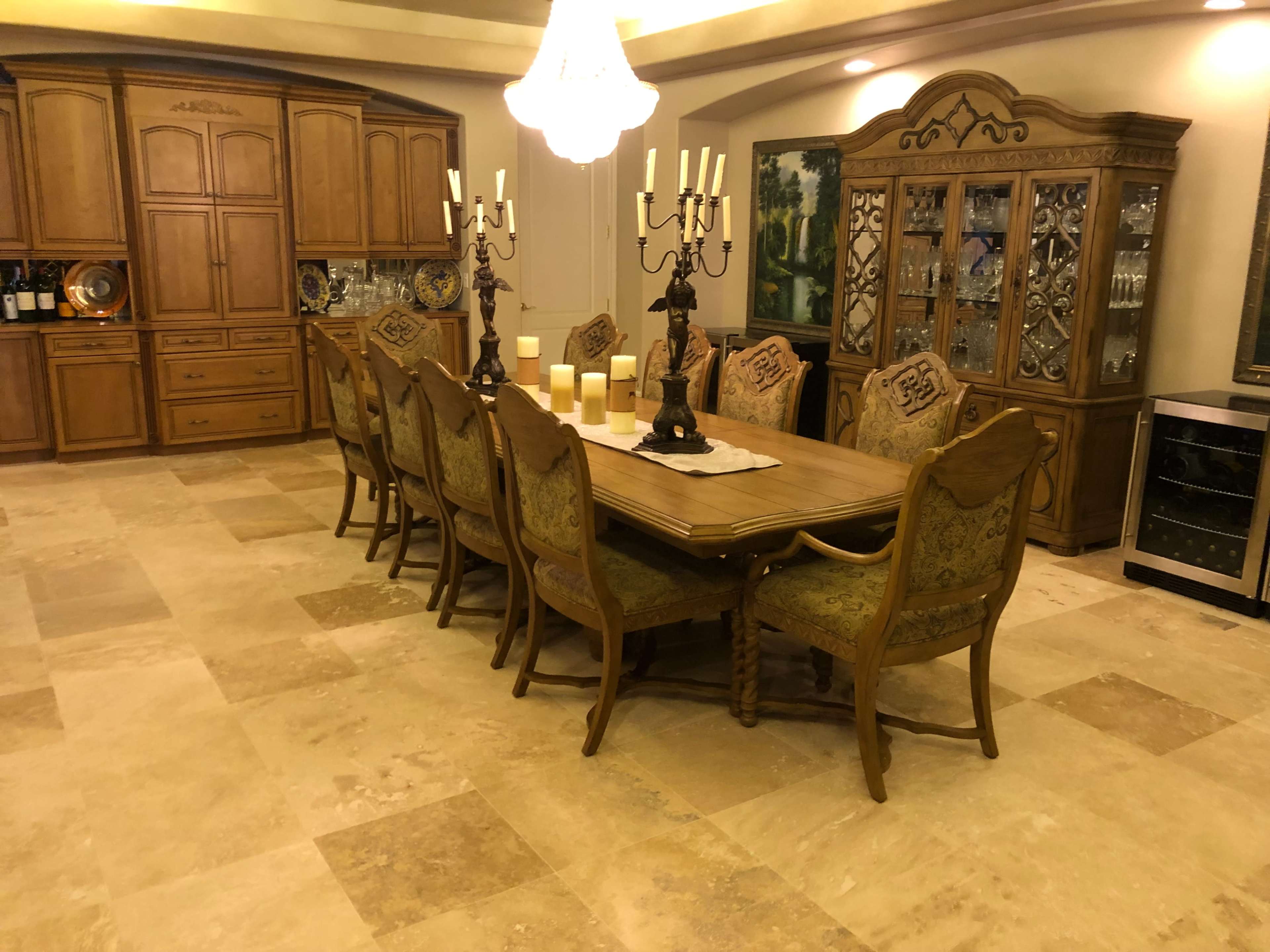 A large wooden dining table surrounded by upholstered chairs is set in a spacious dining area with stone tile flooring and a chandelier above.