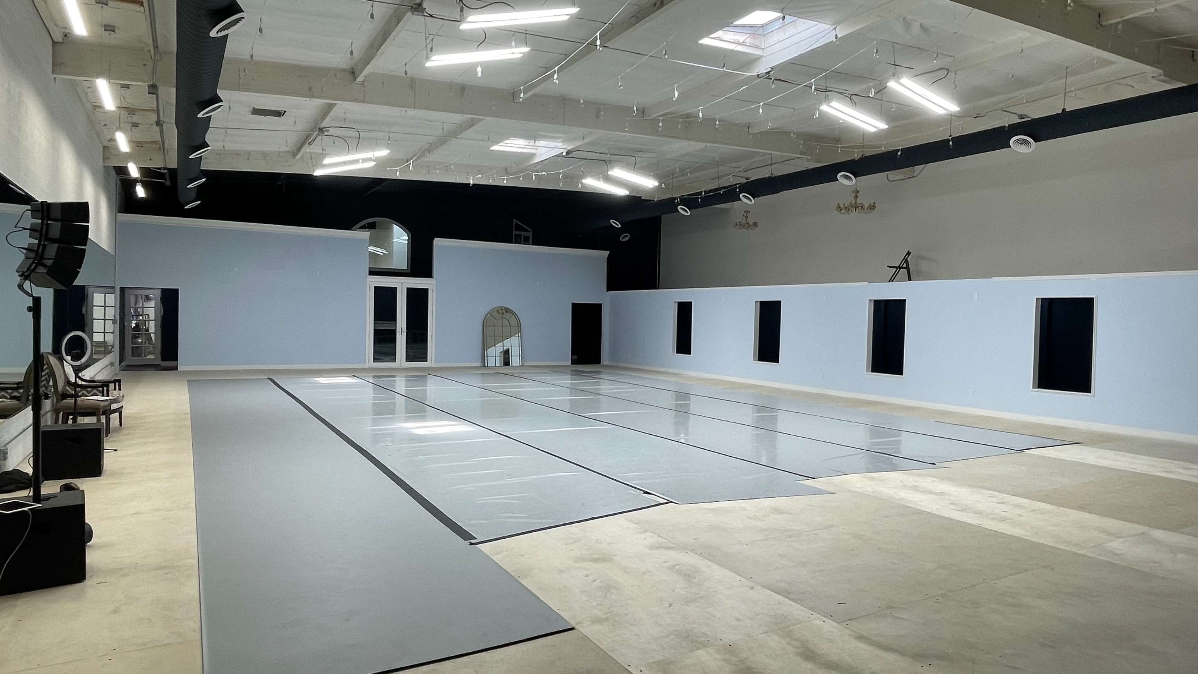 A spacious, well-lit room with empty gray flooring and light blue walls, featuring several windows and a large open area.