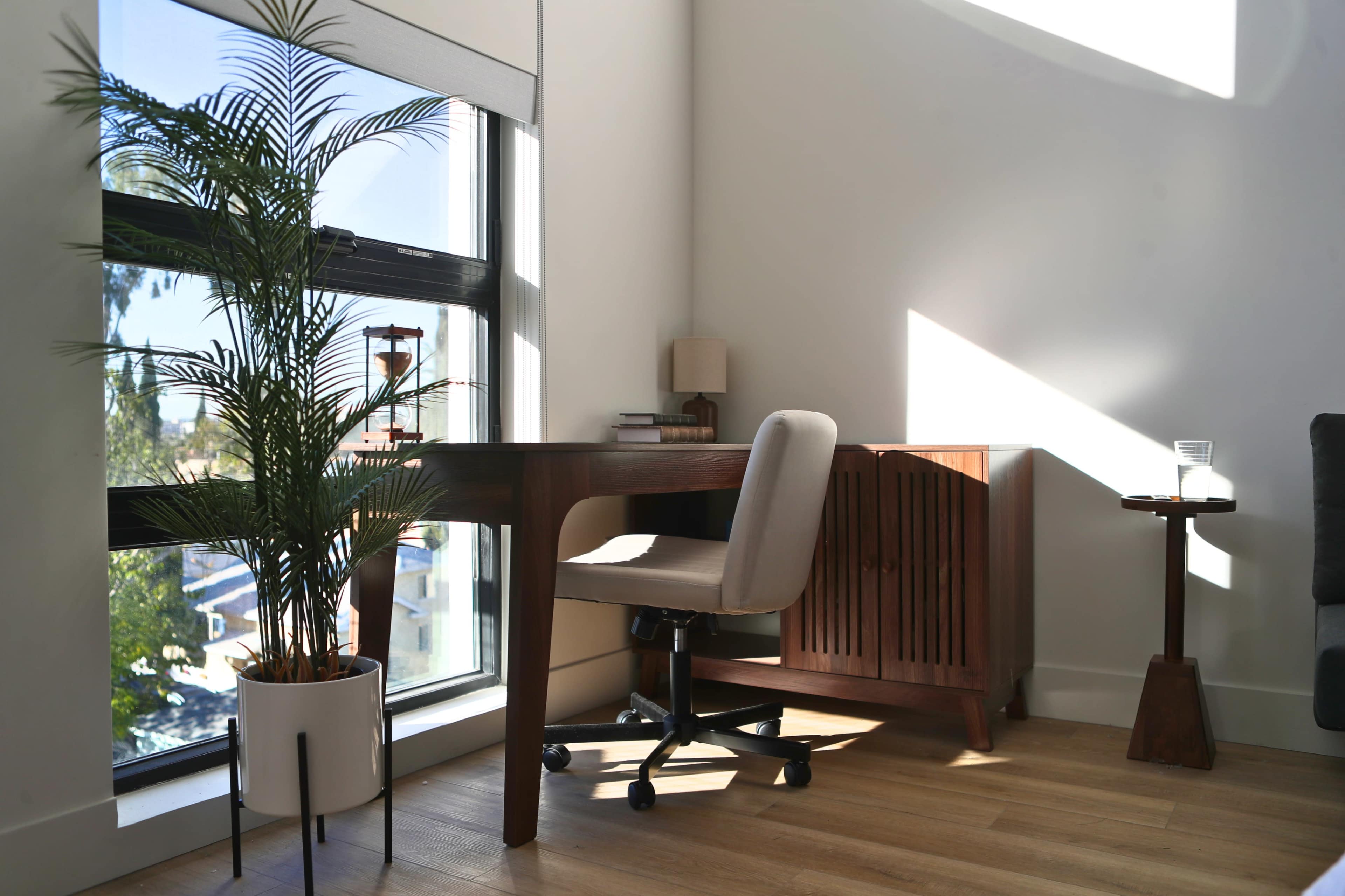 The image shows a modern office space featuring a wooden desk with a chair, a plant in a pot, and a side cabinet, all illuminated by natural light coming through a large window.