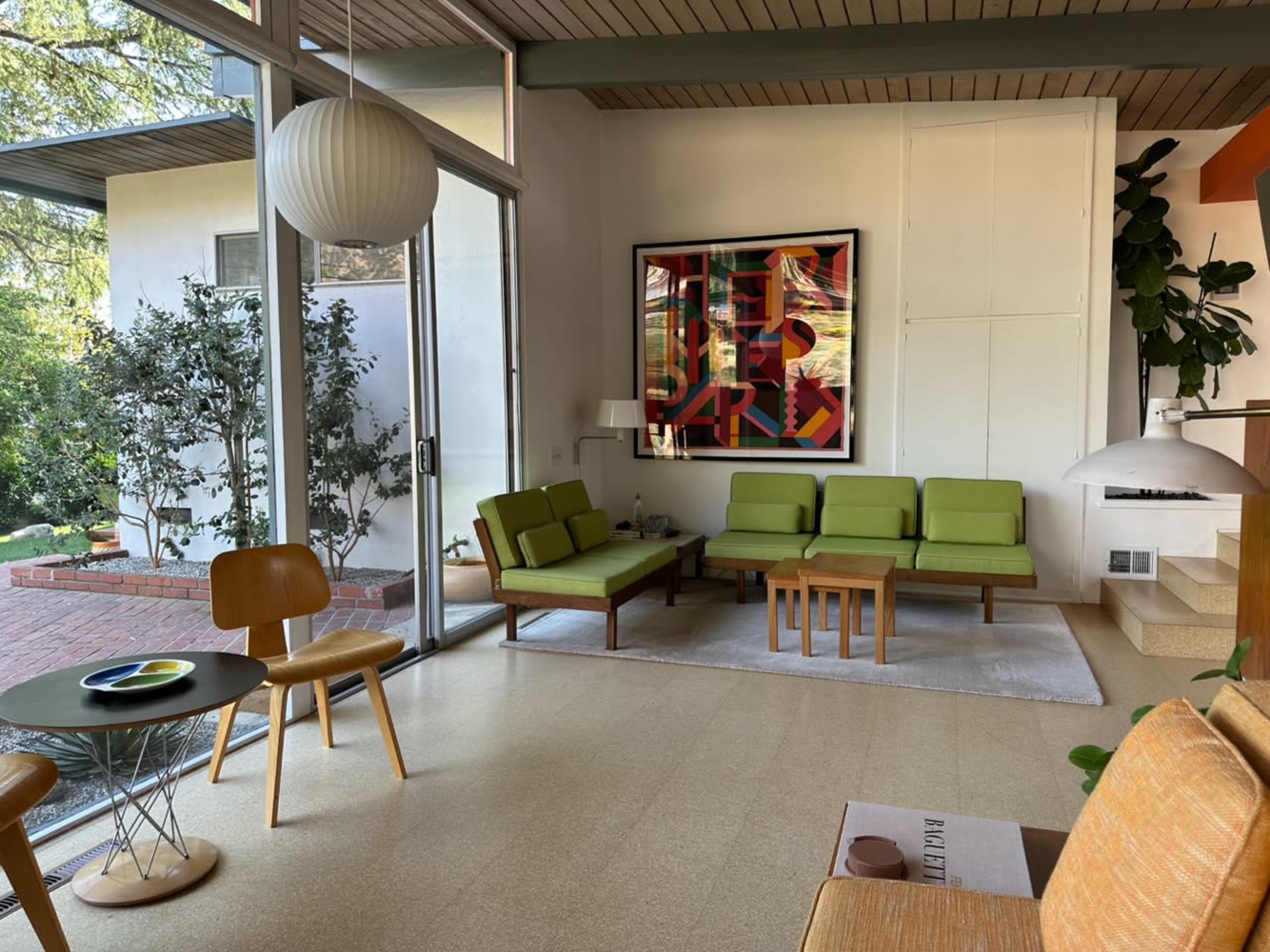 The scene features a modern living room with green sofas, a wooden coffee table, and a large framed artwork, all illuminated by natural light from substantial windows.