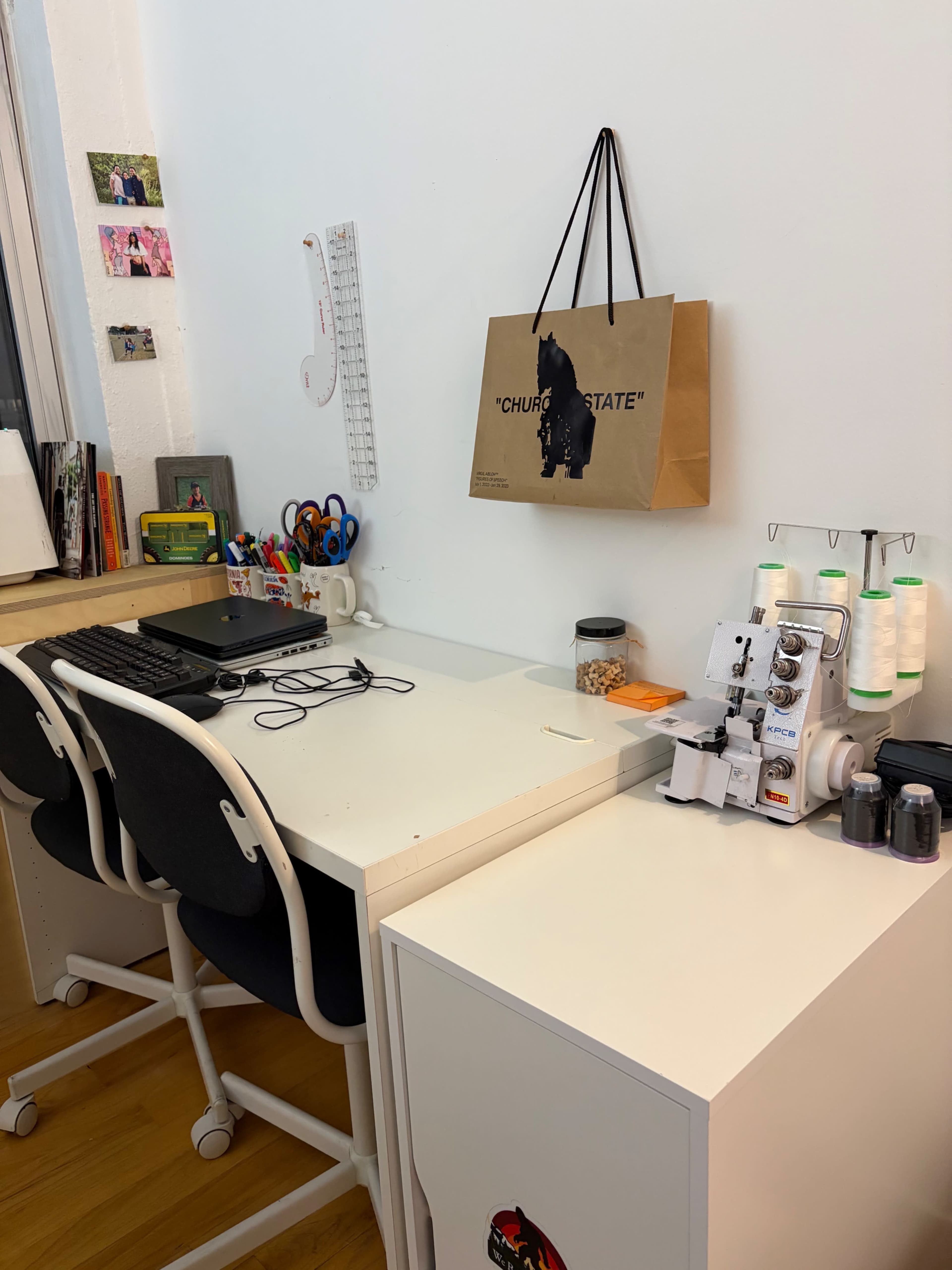 The image shows a clean, organized workspace featuring two chairs at a white desk with various office supplies, a decorative bag hanging on the wall, and a small cabinet beside the desk.