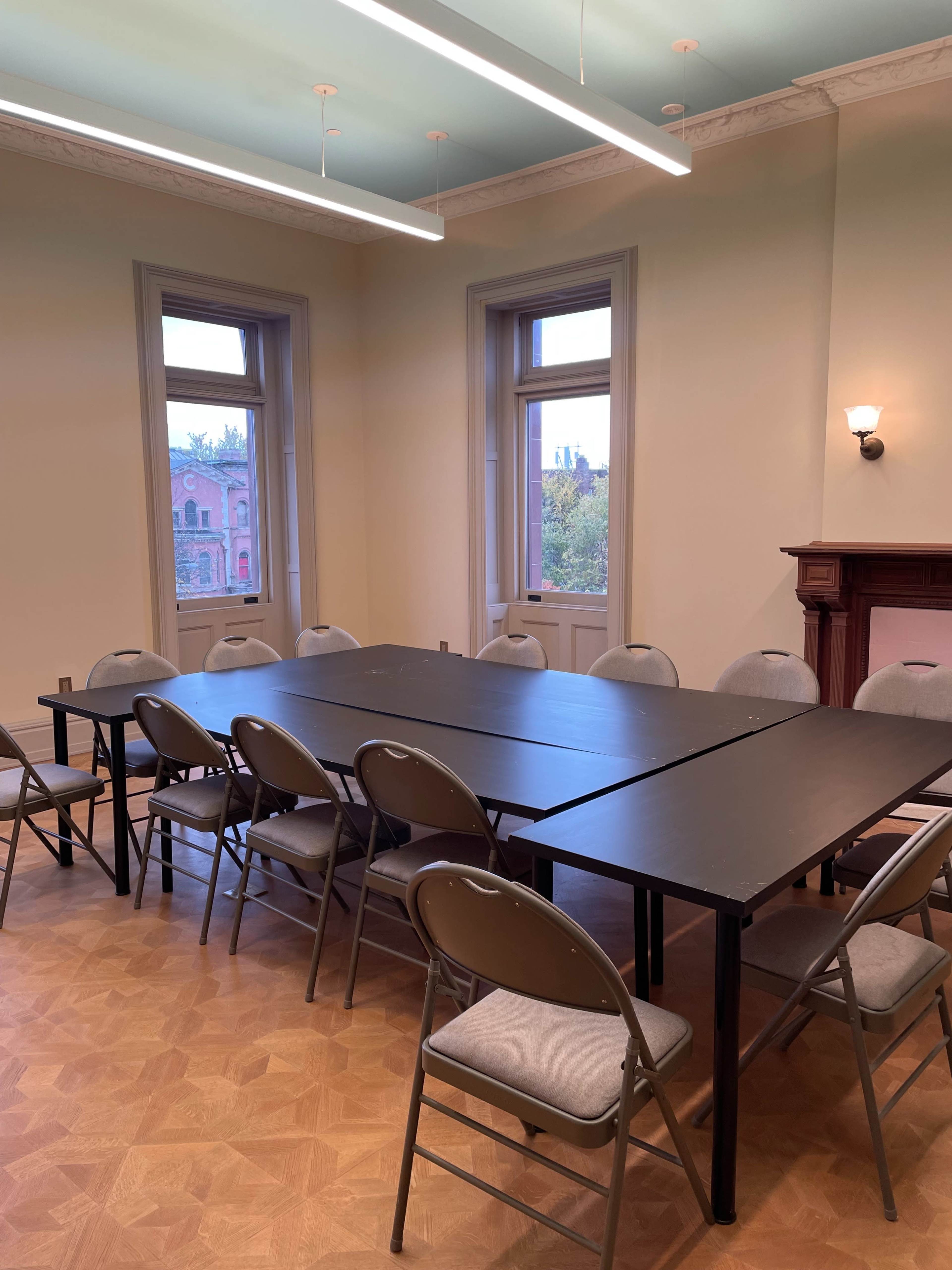 The image shows a meeting room with a black rectangular table surrounded by gray folding chairs, illuminated by natural light from two windows.