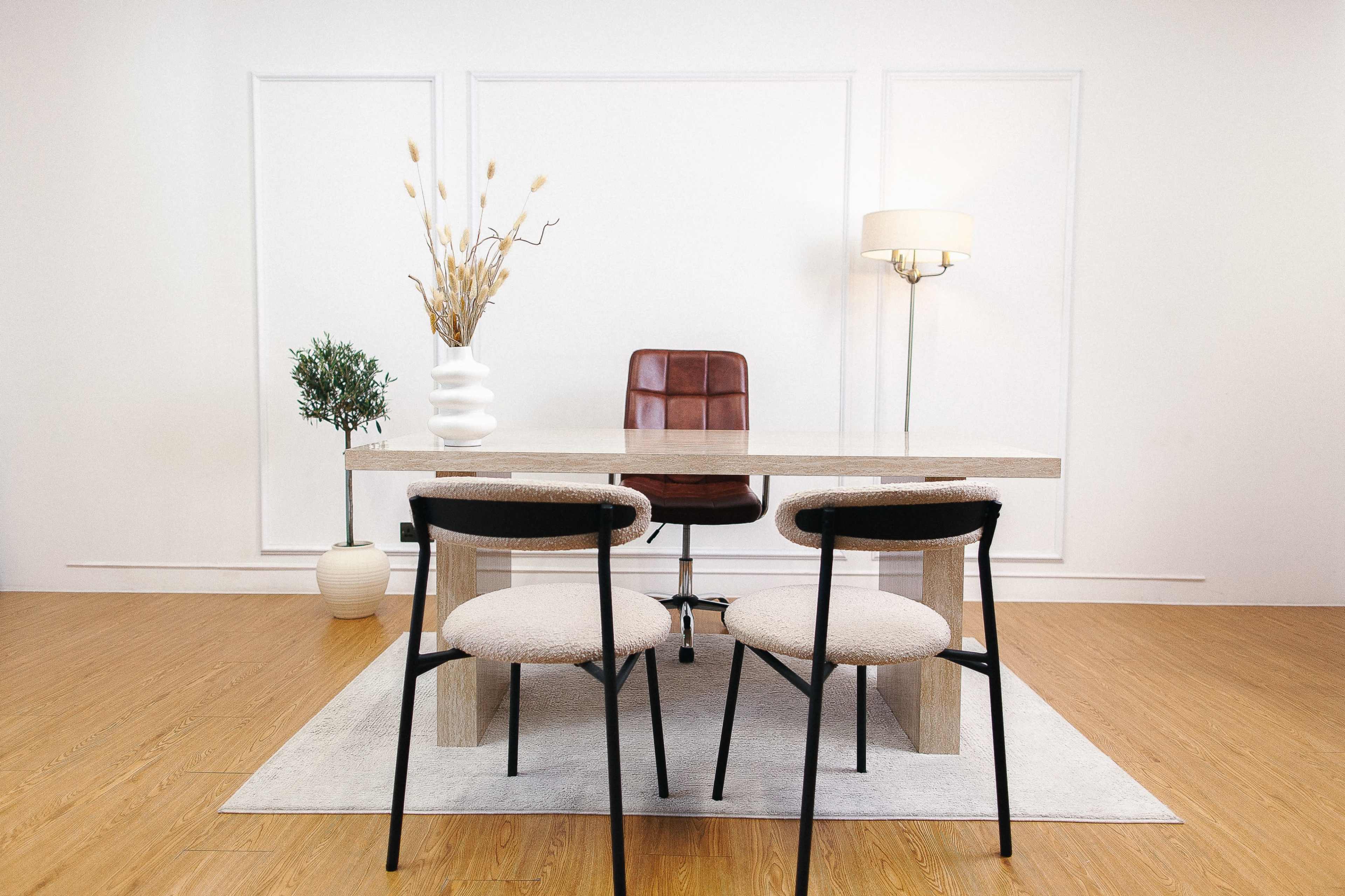 A modern office space features a wooden table with two upholstered chairs, a brown leather office chair, a vase with dried plants, and a small potted tree against a white backdrop.
