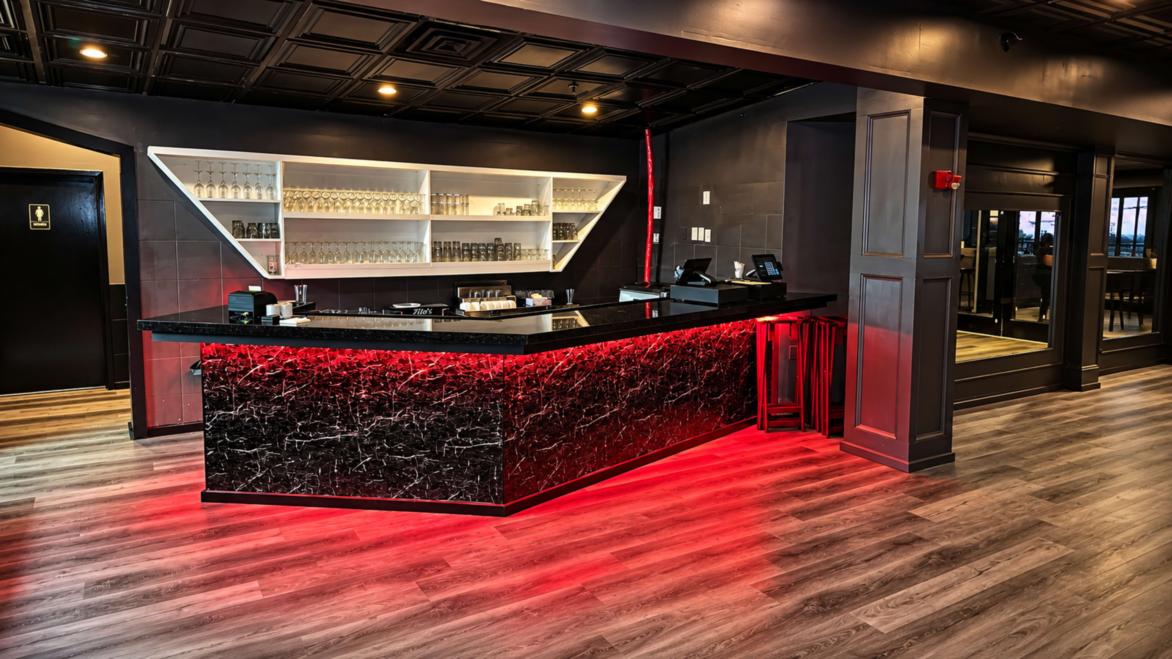 The image shows a modern bar area featuring a sleek black countertop, illuminated by red lighting, with shelves displaying glassware in the background.
