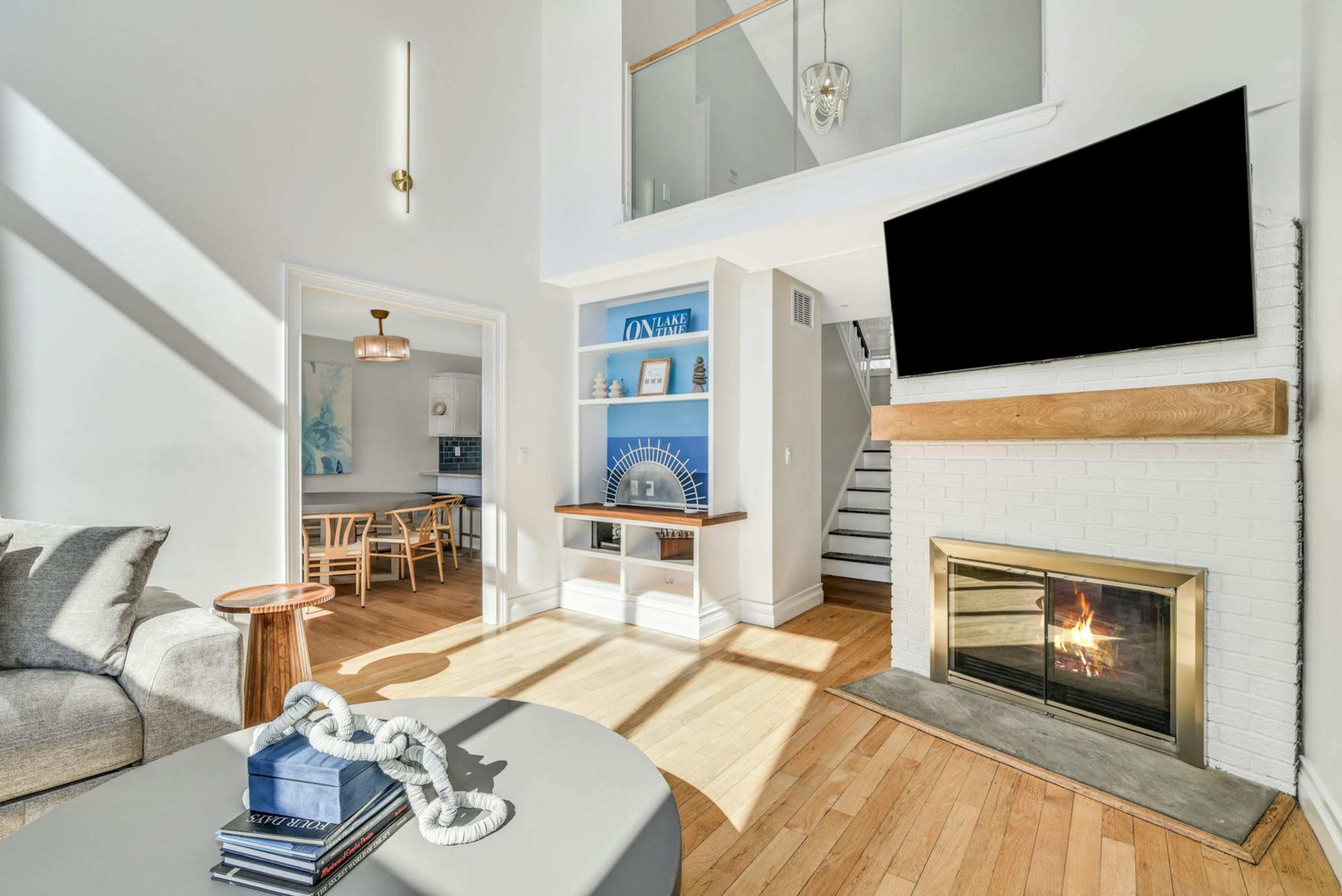 The image shows a bright living room with a fireplace, a large television mounted on the wall, and a dining area visible in the background.