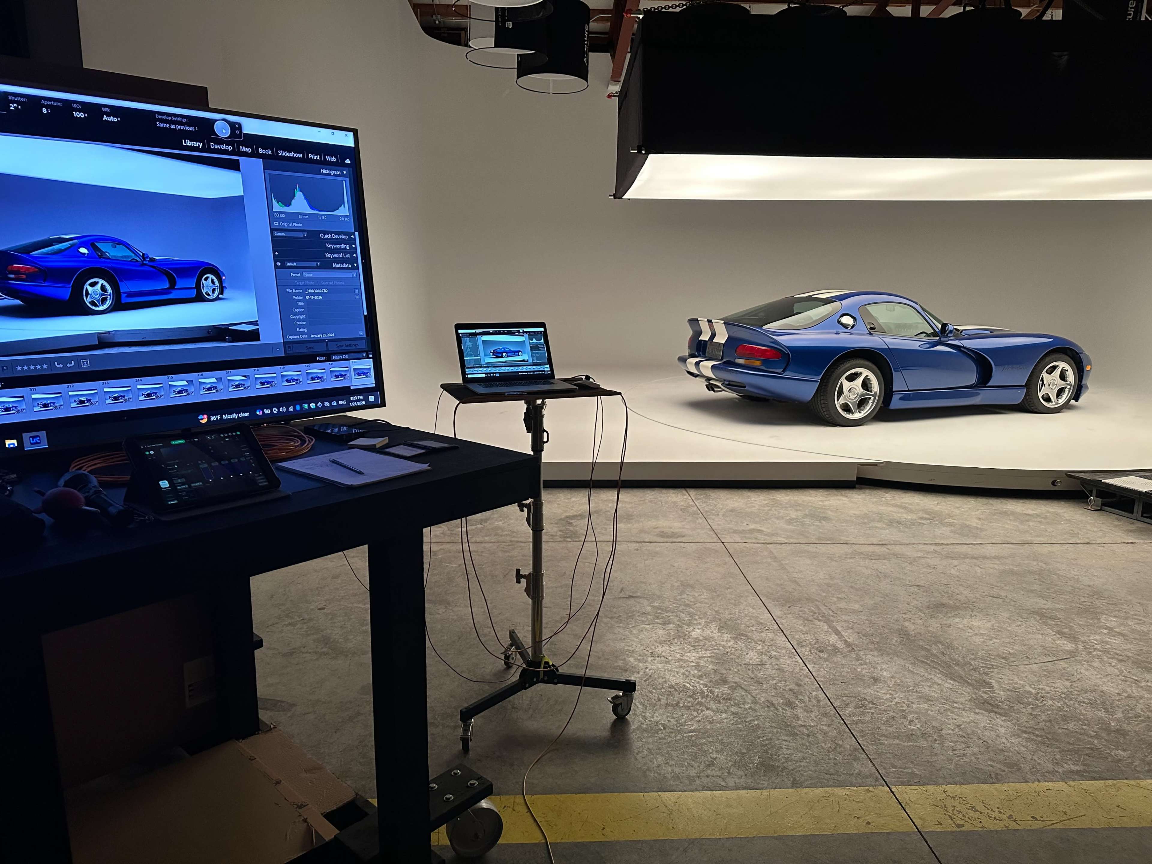 A blue sports car is positioned on a neutral backdrop in a photo studio, with a computer setup displaying an image of the car on a monitor.