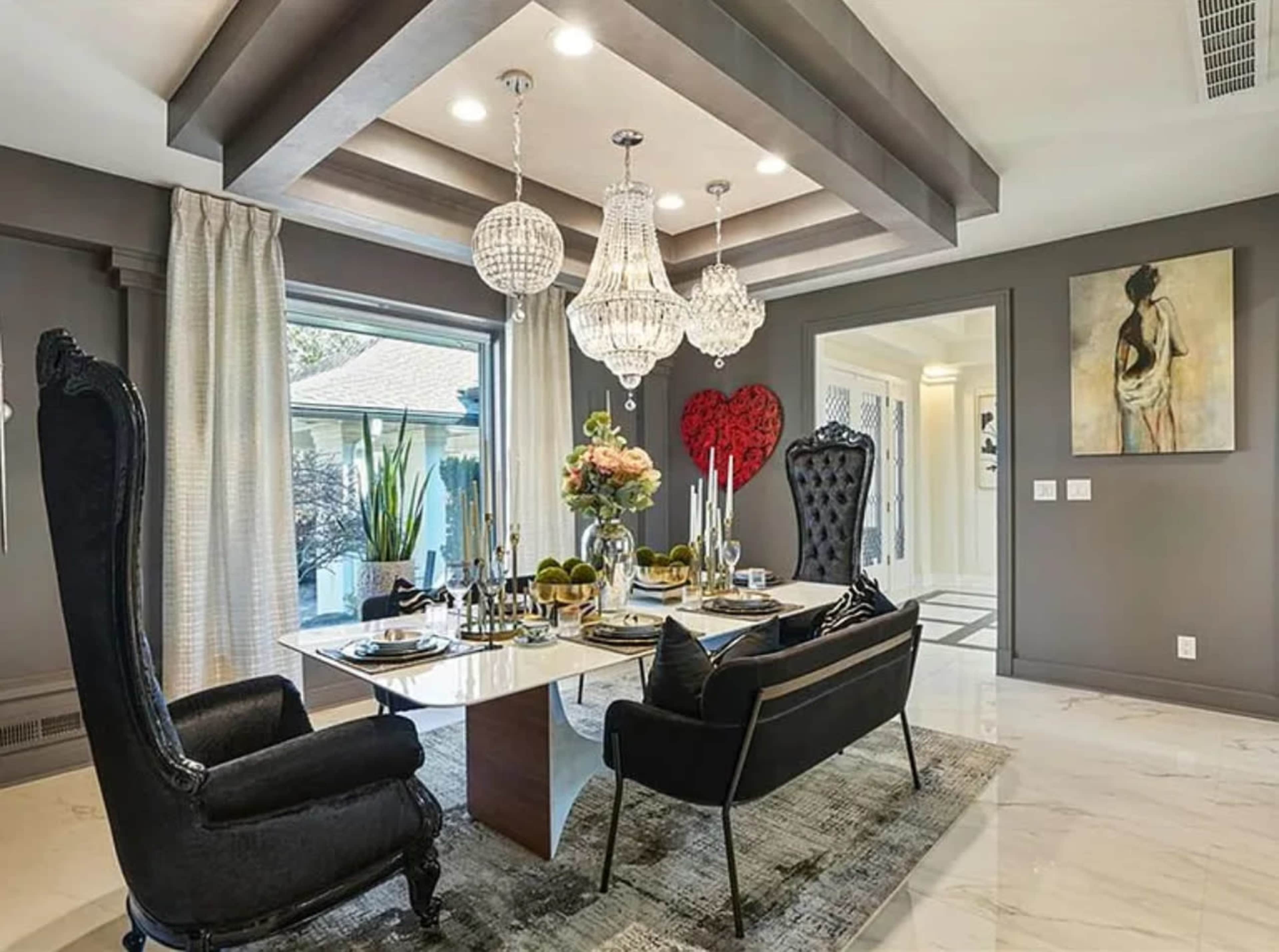 The image shows a modern dining room featuring a table set for a meal, two large black chairs, a floral centerpiece, and decorative lighting.
