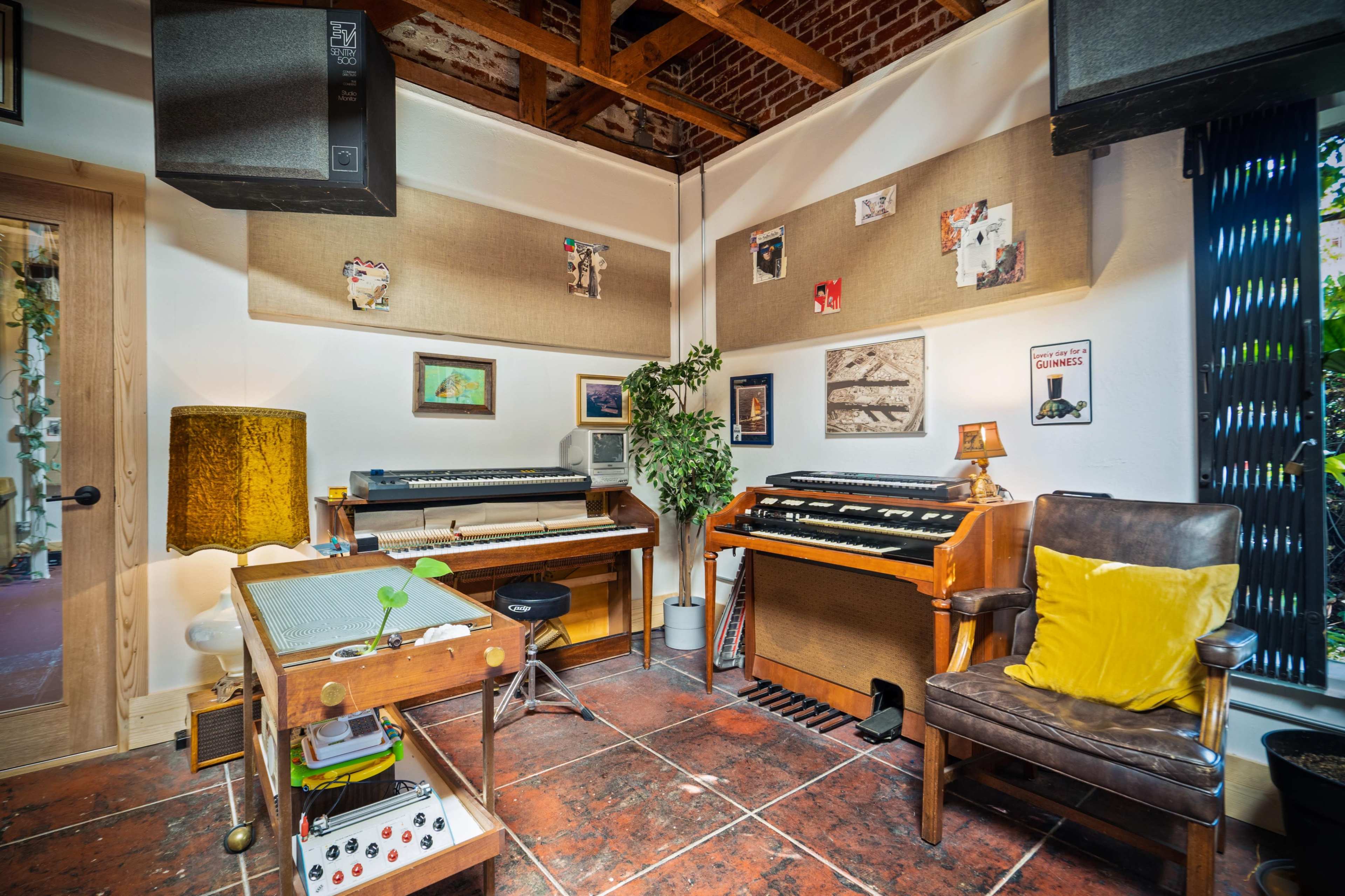 A music studio with various electronic keyboards, a wooden desk, and a cozy chair, all set against a backdrop of exposed brick and wooden beams.