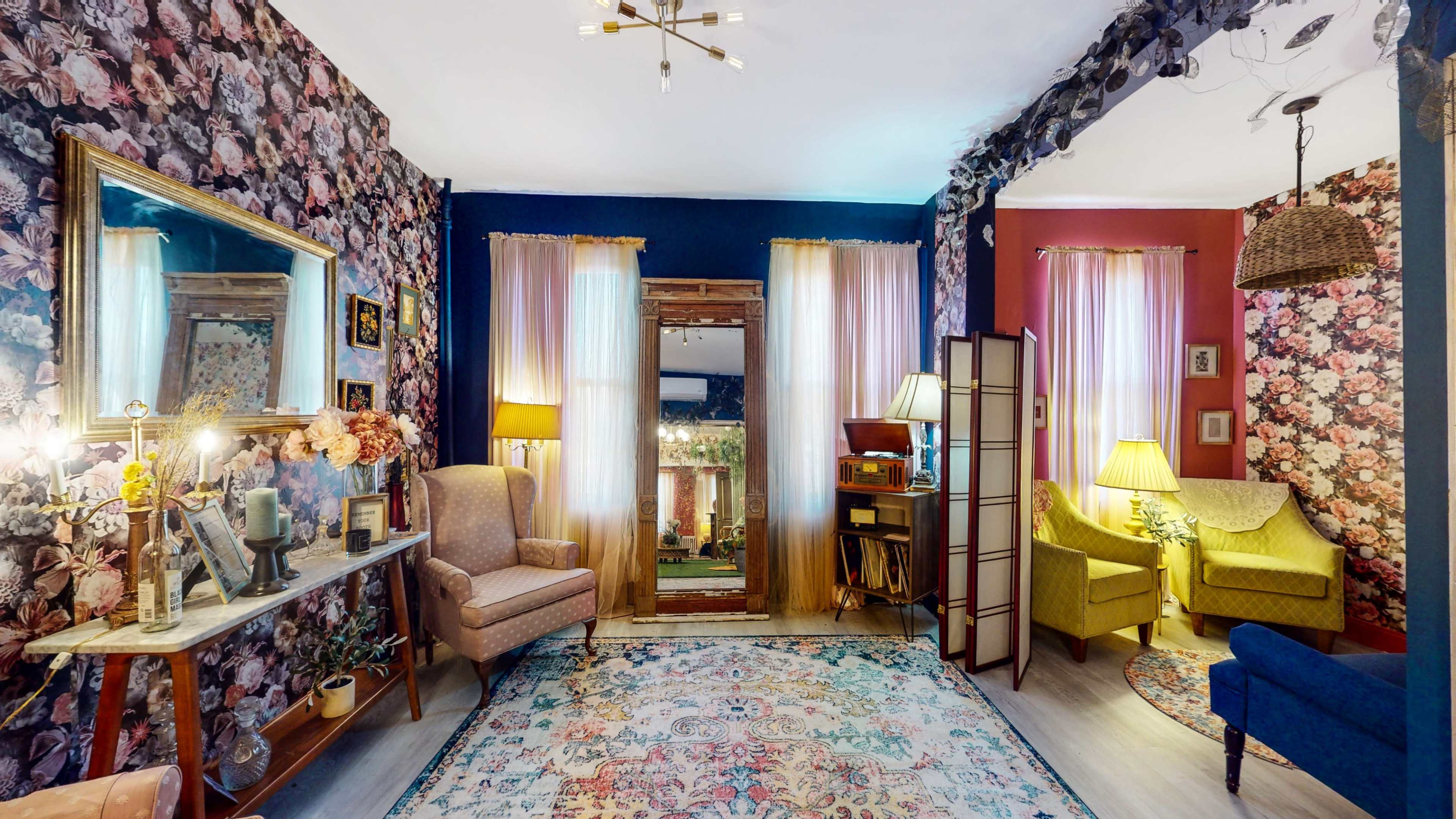 A living room decorated with floral wallpaper, featuring a seating area with two chairs and a sofa, a decorative mirror, and a doorway leading to an outdoor space.