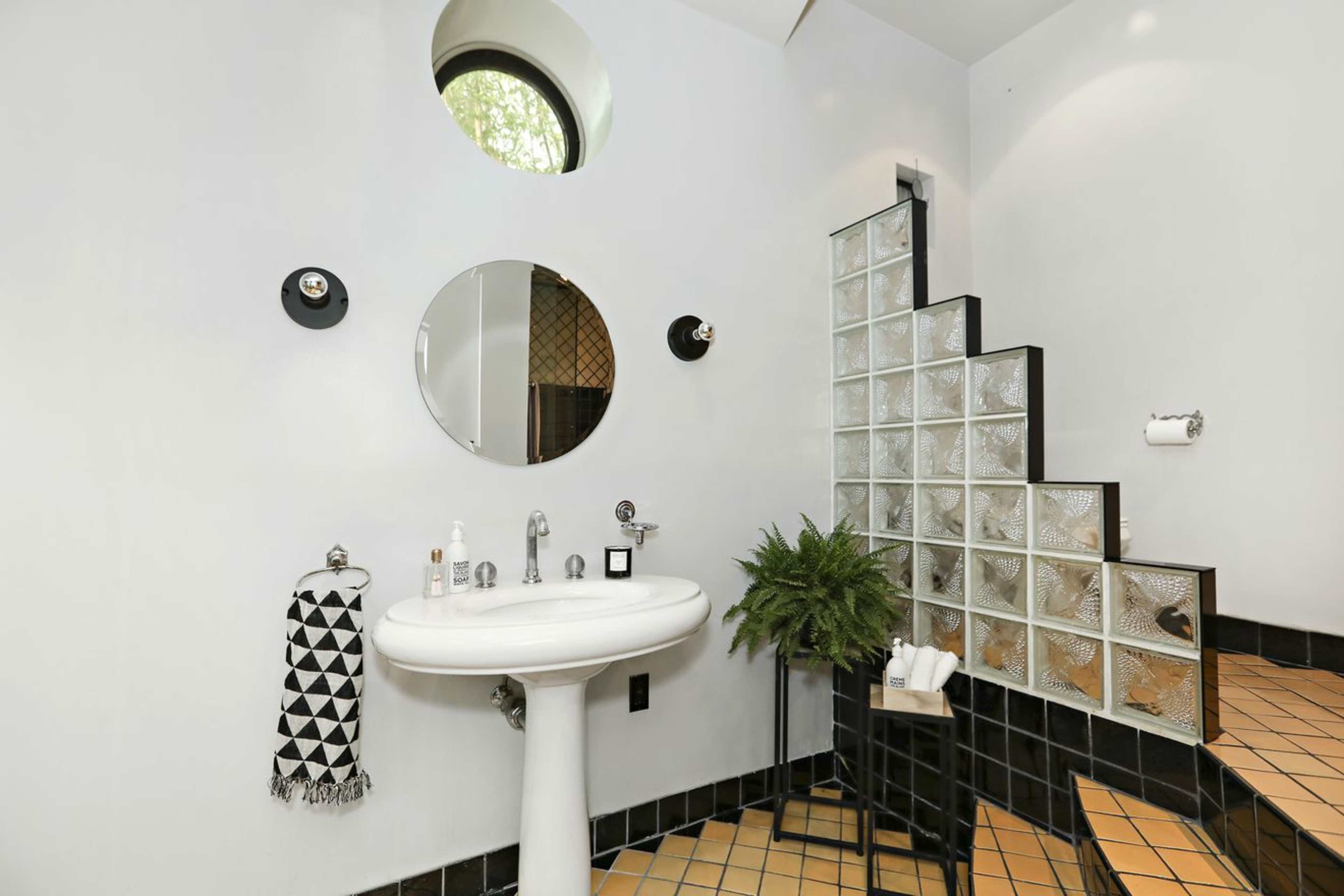 The image shows a modern bathroom with a round mirror, a pedestal sink, glass block walls, and a staircase leading upward.