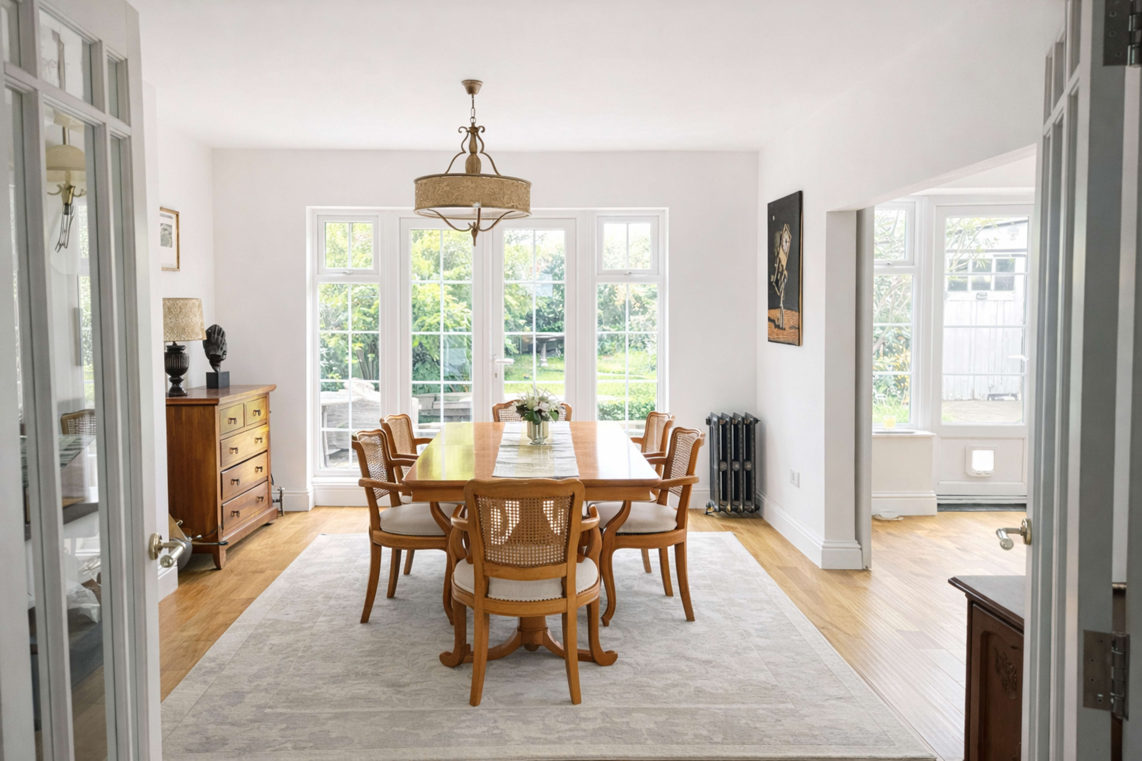 A bright dining room features a wooden table surrounded by eight chairs, with large windows allowing natural light to illuminate the space.