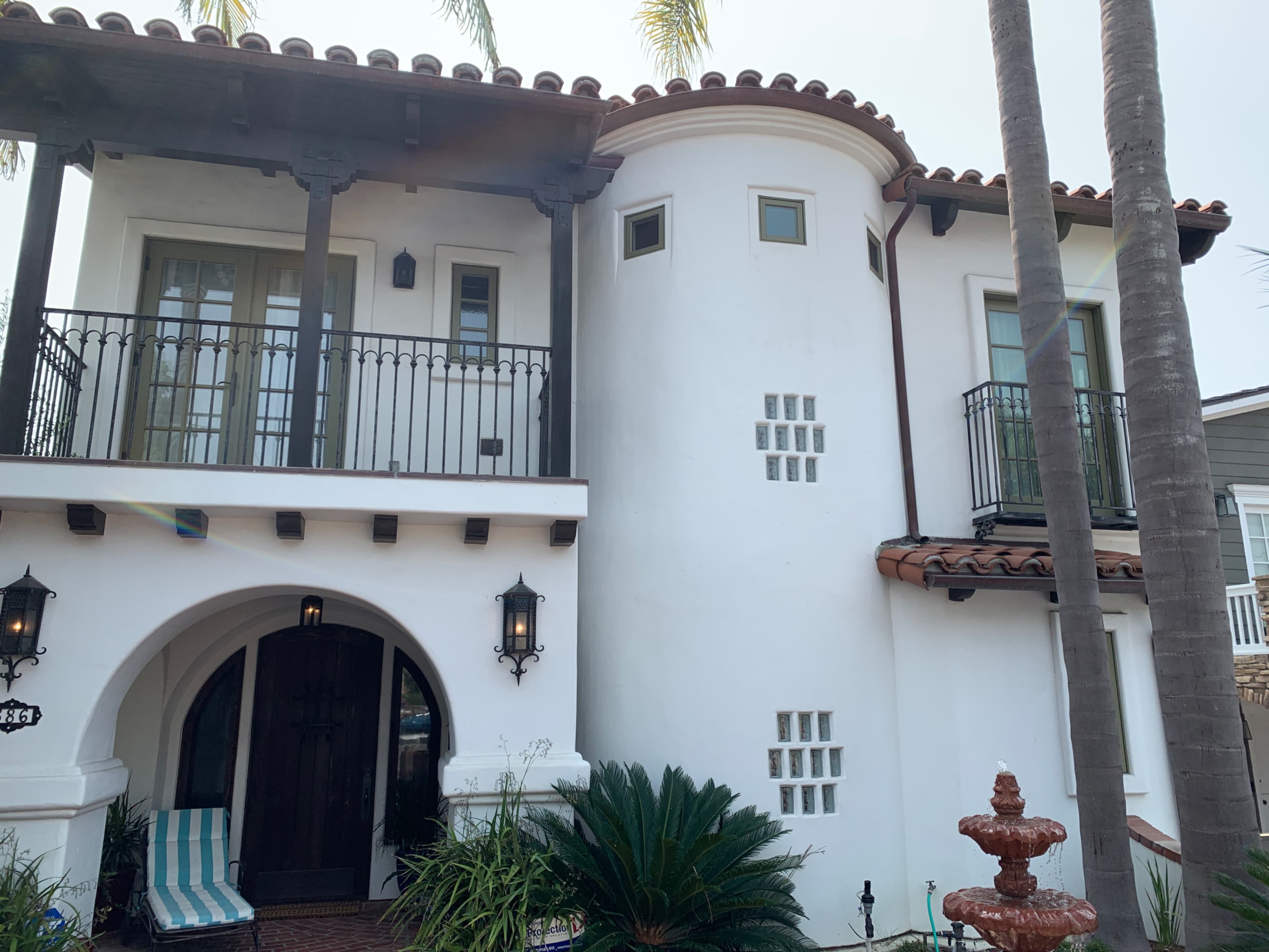 A two-story white stucco house with a rounded corner and balconies, flanked by palm trees and featuring a front entrance with a wooden door.