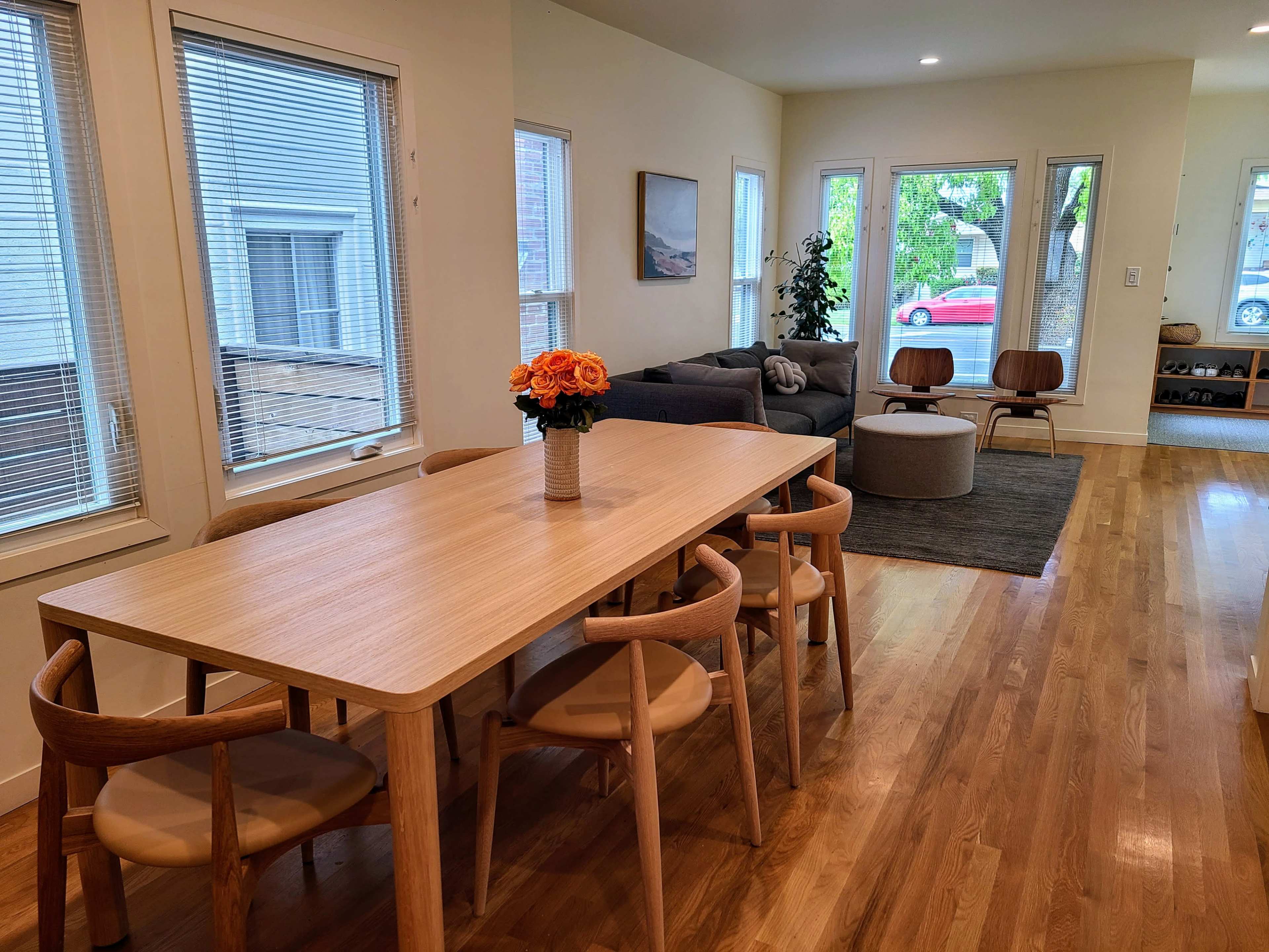 A well-lit dining area features a wooden table with six chairs, a vase of flowers in the center, and an adjacent living room with a sofa and two chairs.