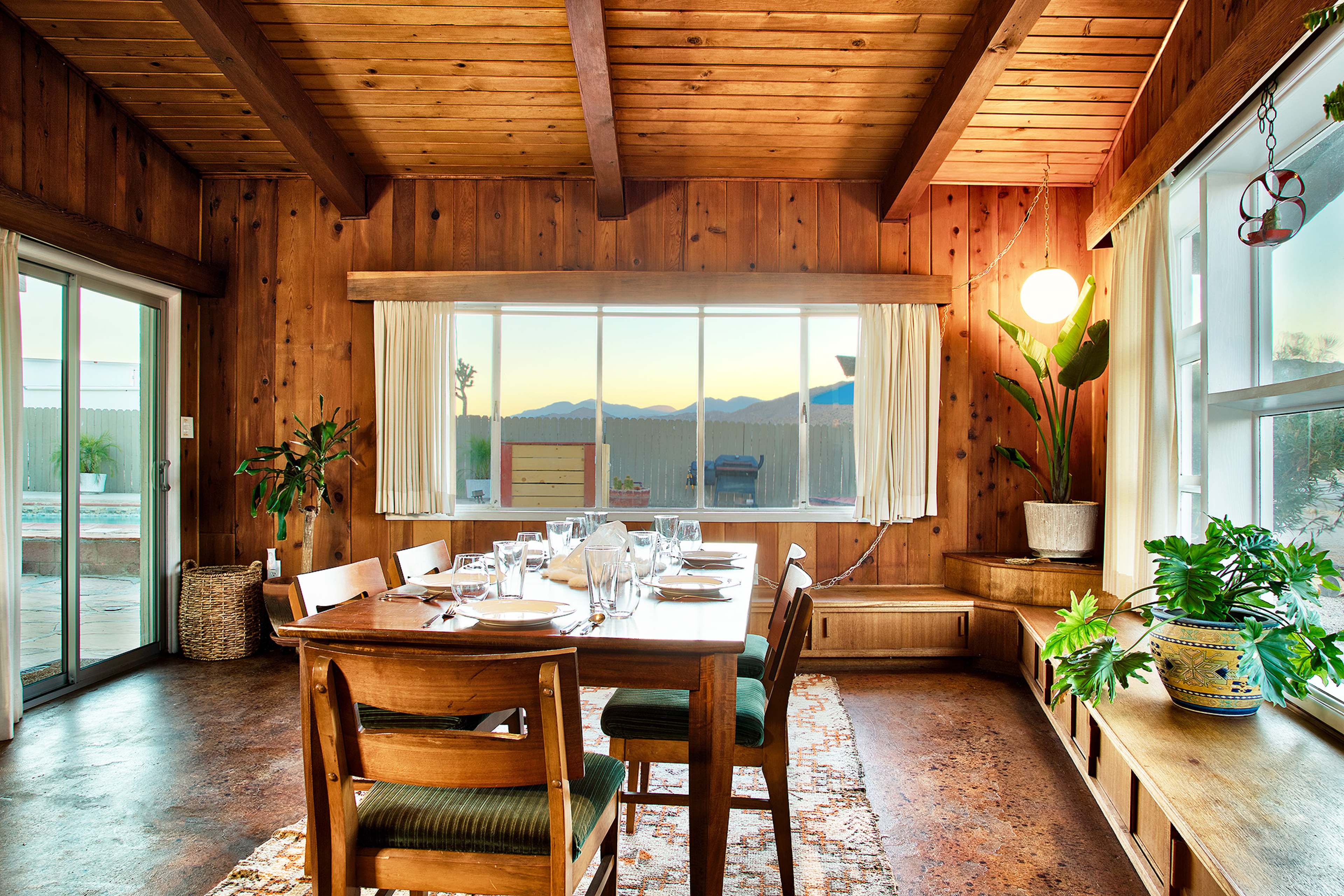 A wooden dining room features a table set for a meal, with large windows displaying mountains in the background.