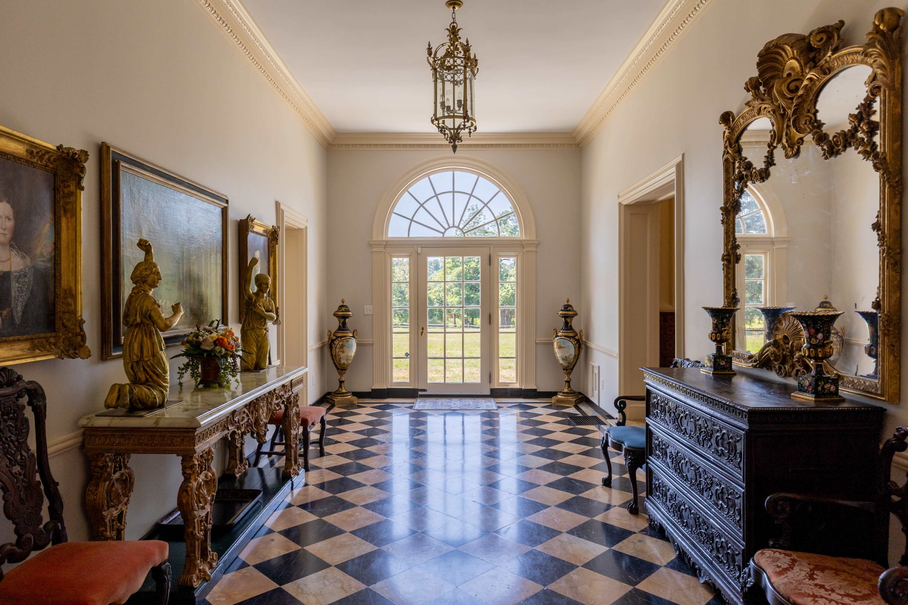 The image shows a spacious hallway with a checkered floor, featuring large windows, ornate mirrors, and decorative furniture.