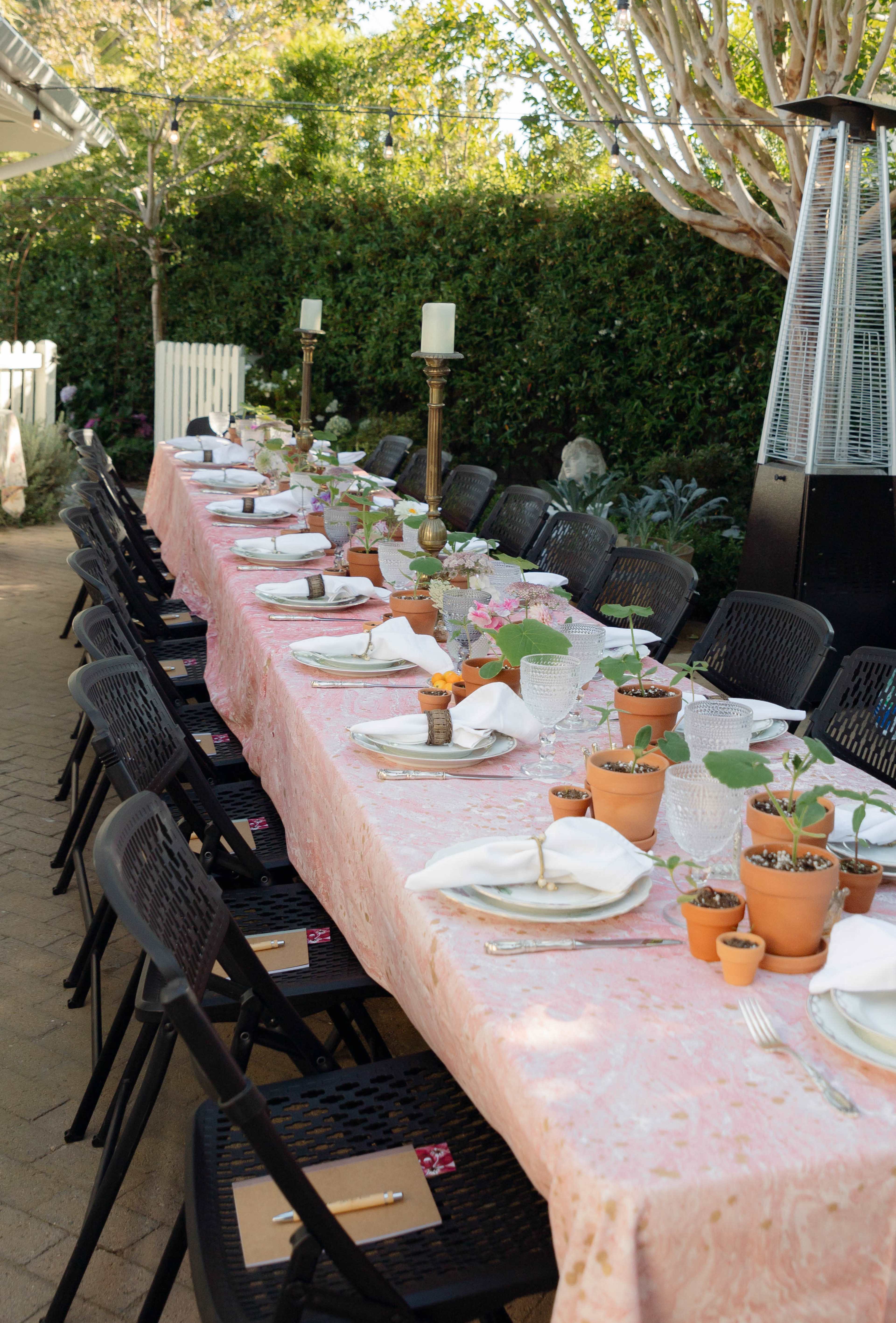 A long outdoor dining table is set with white plates, folded napkins, and small potted plants as centerpieces, surrounded by black chairs.