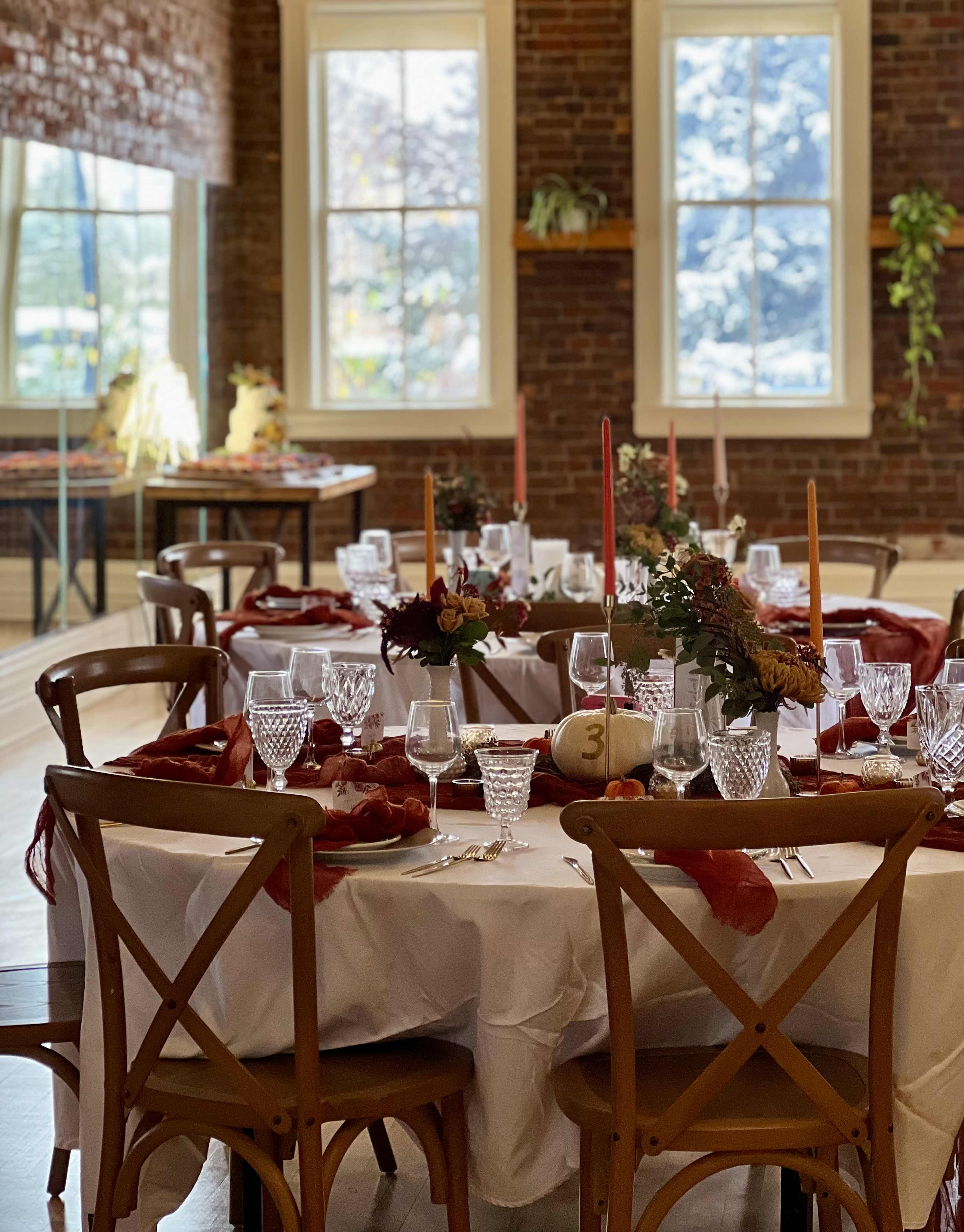 A round dining table is elegantly set with glassware, cutlery, and floral centerpieces, surrounded by wooden chairs in a warmly lit room with brick walls and large windows.