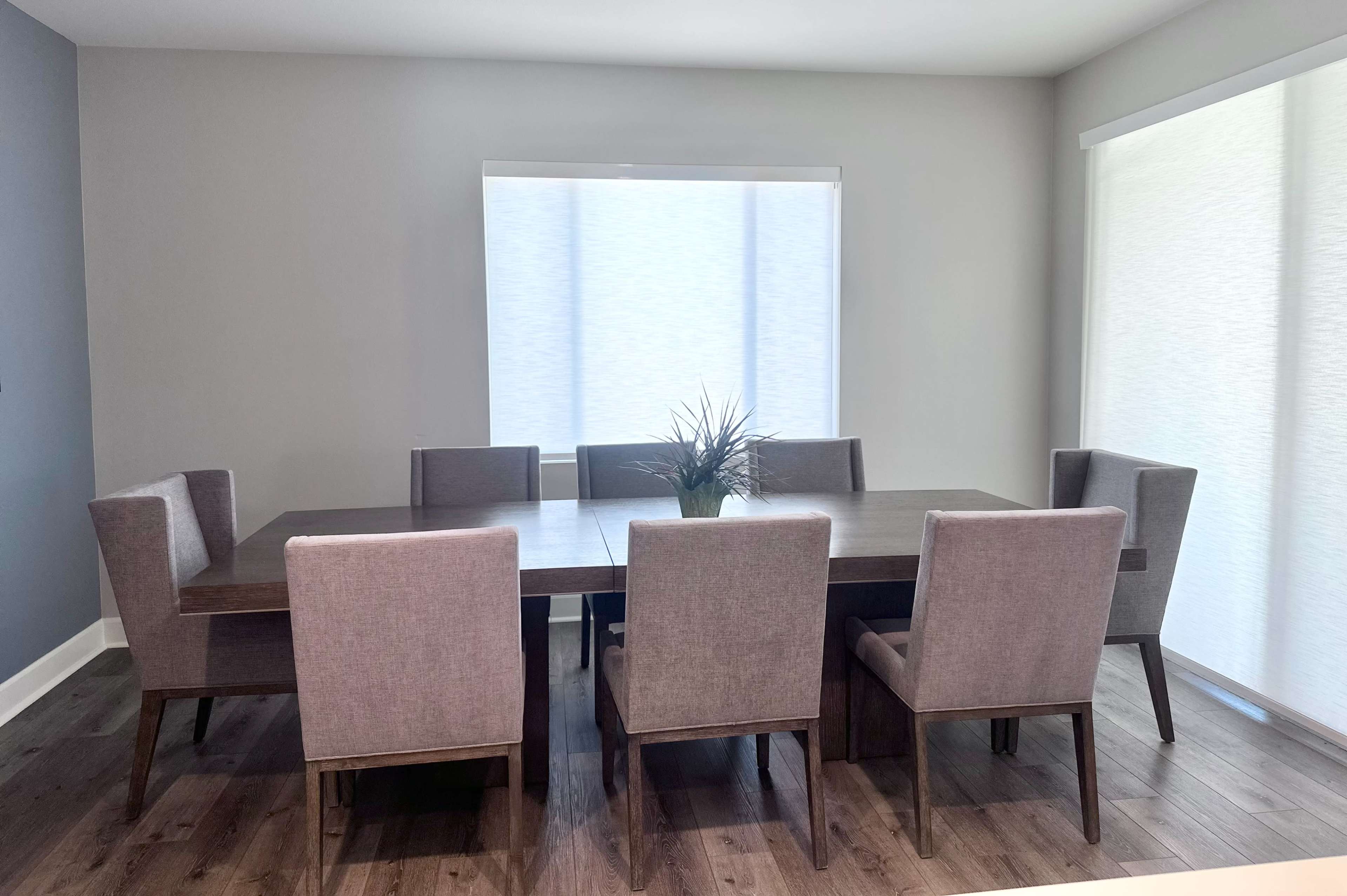 A long wooden dining table is surrounded by six upholstered chairs in a room with large windows and light-colored walls.