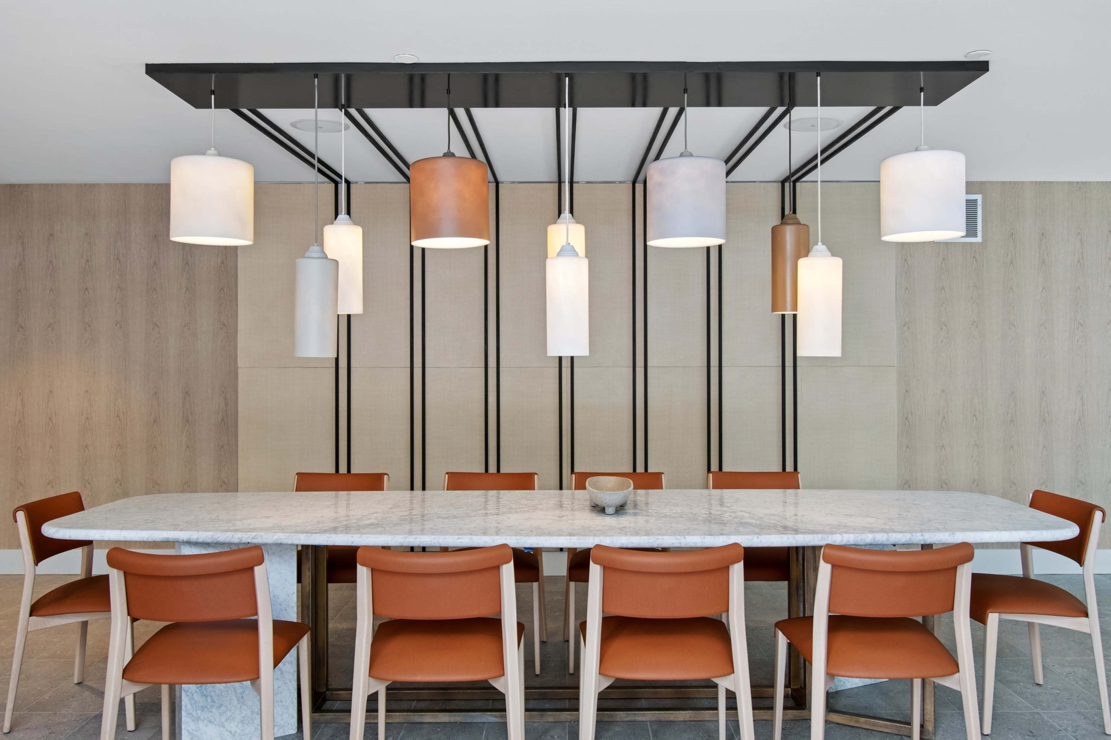 The image shows a modern dining table with twelve chairs arranged around it, illuminated by multiple pendant lights hanging from a black fixture.