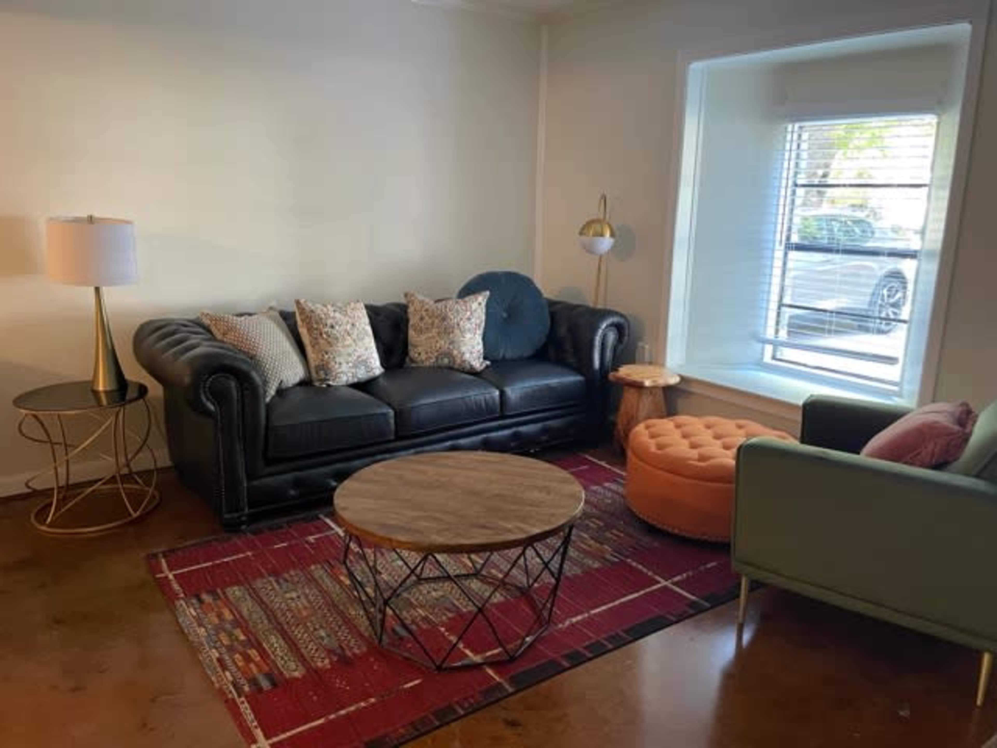 A living room with a black leather sofa, a round wooden coffee table, an orange ottoman, and a window with blinds.