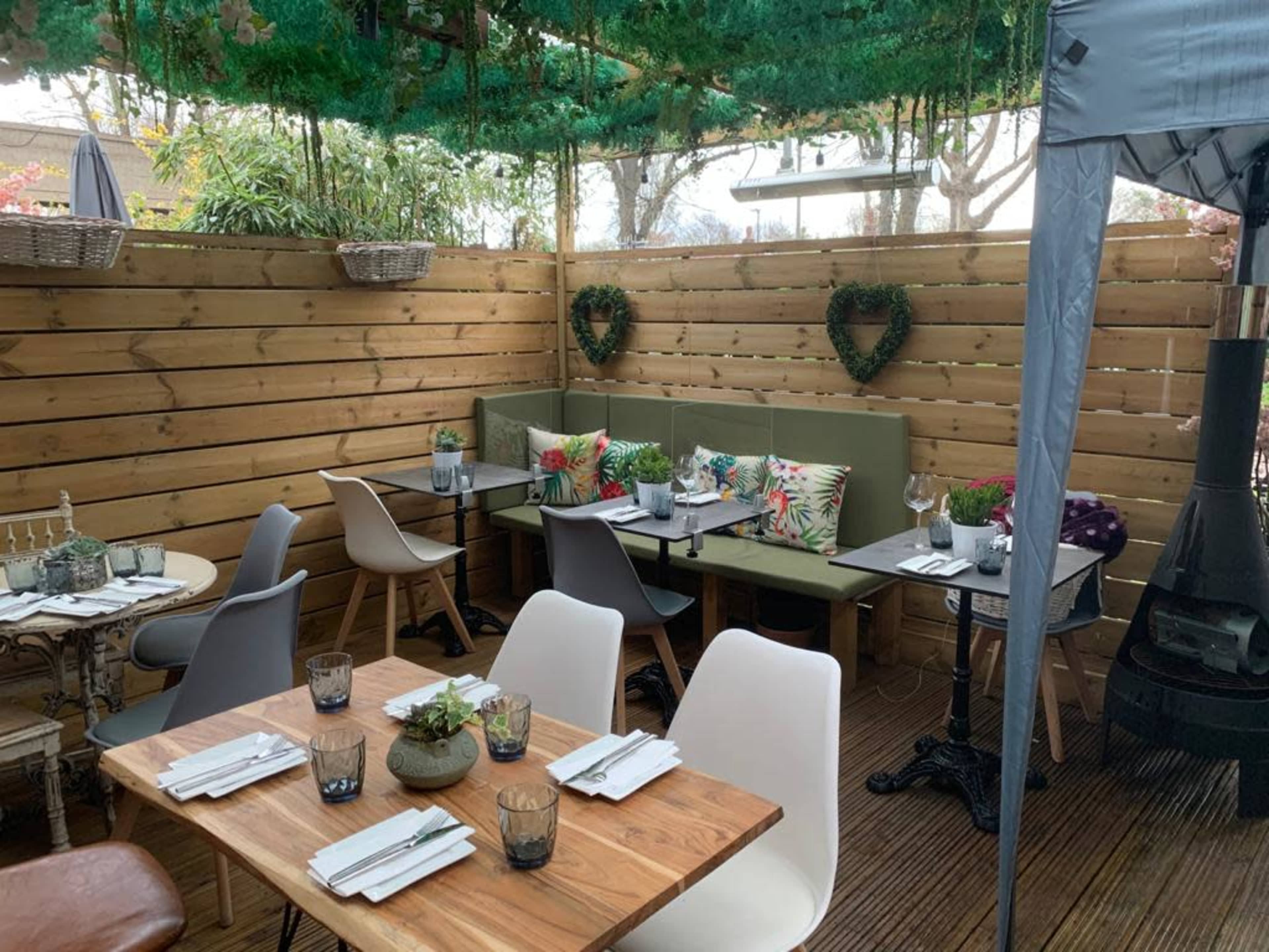 A cozy outdoor dining area with wooden walls, various tables, and seating arrangements adorned with plants and cushions.