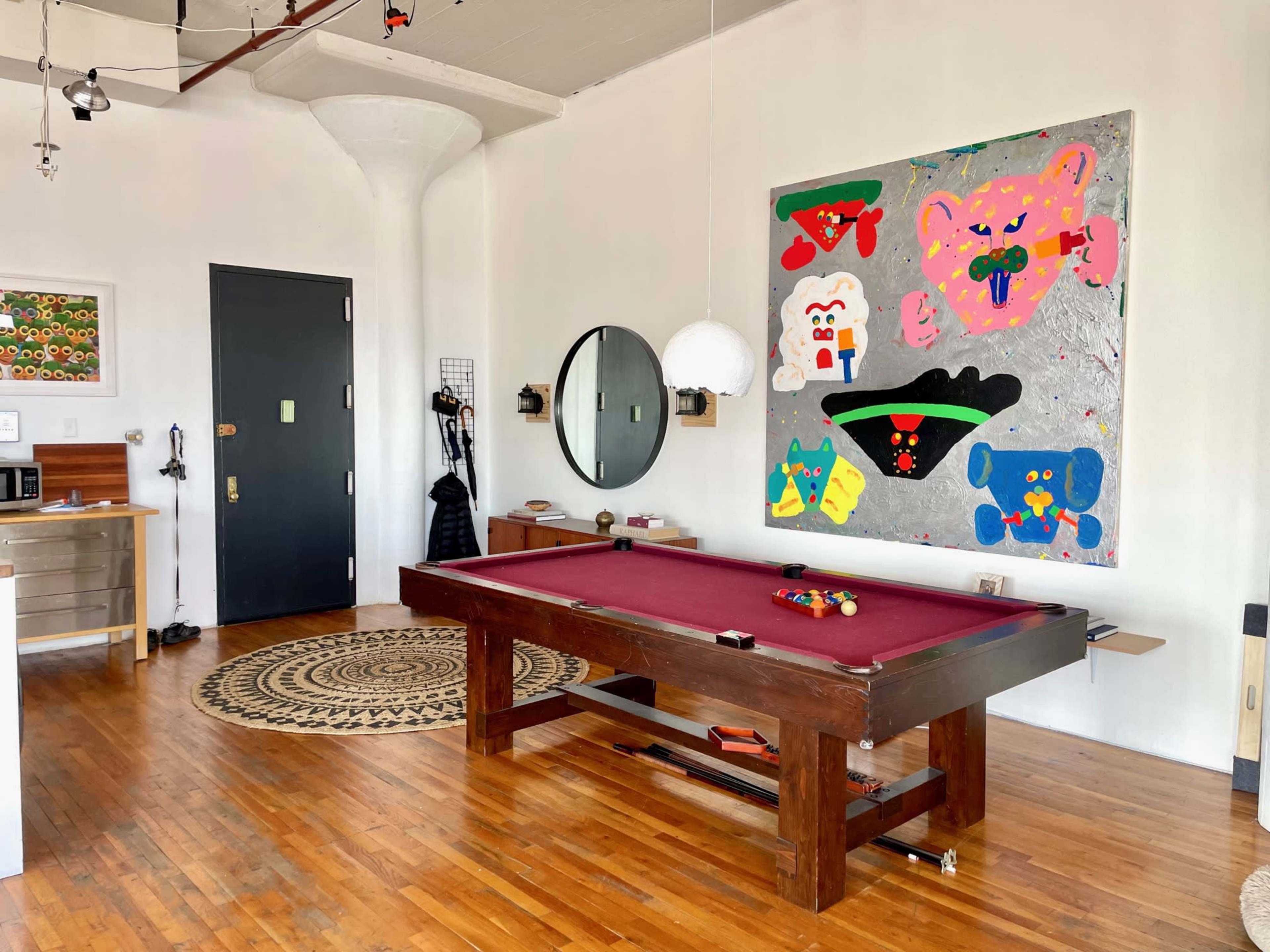 The image shows a room featuring a billiard table, colorful artwork on the wall, and a circular rug on a wooden floor.