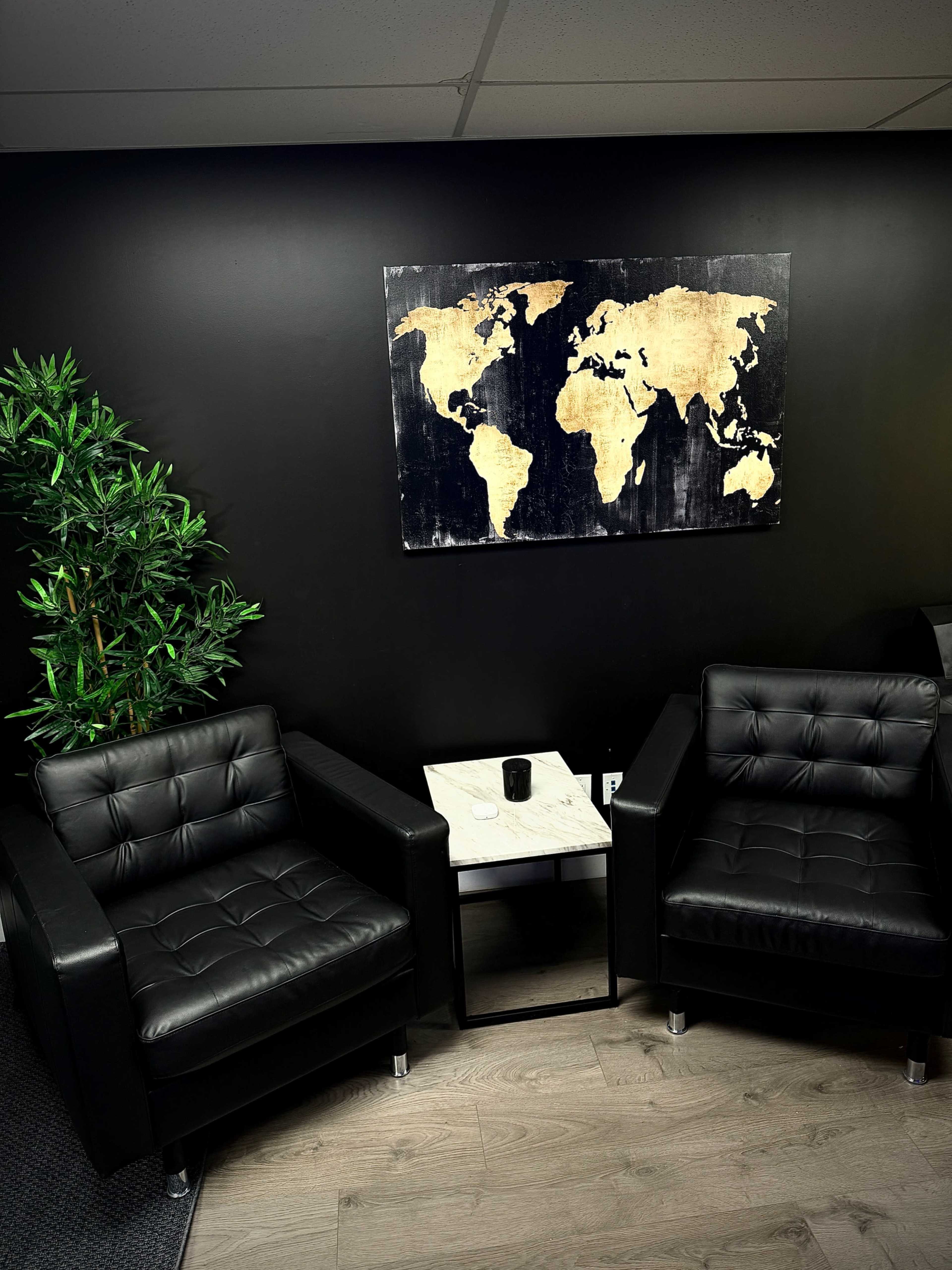 The image shows two black leather armchairs facing a small marble-topped table, with a wooden world map hung on a black wall and a potted plant nearby.