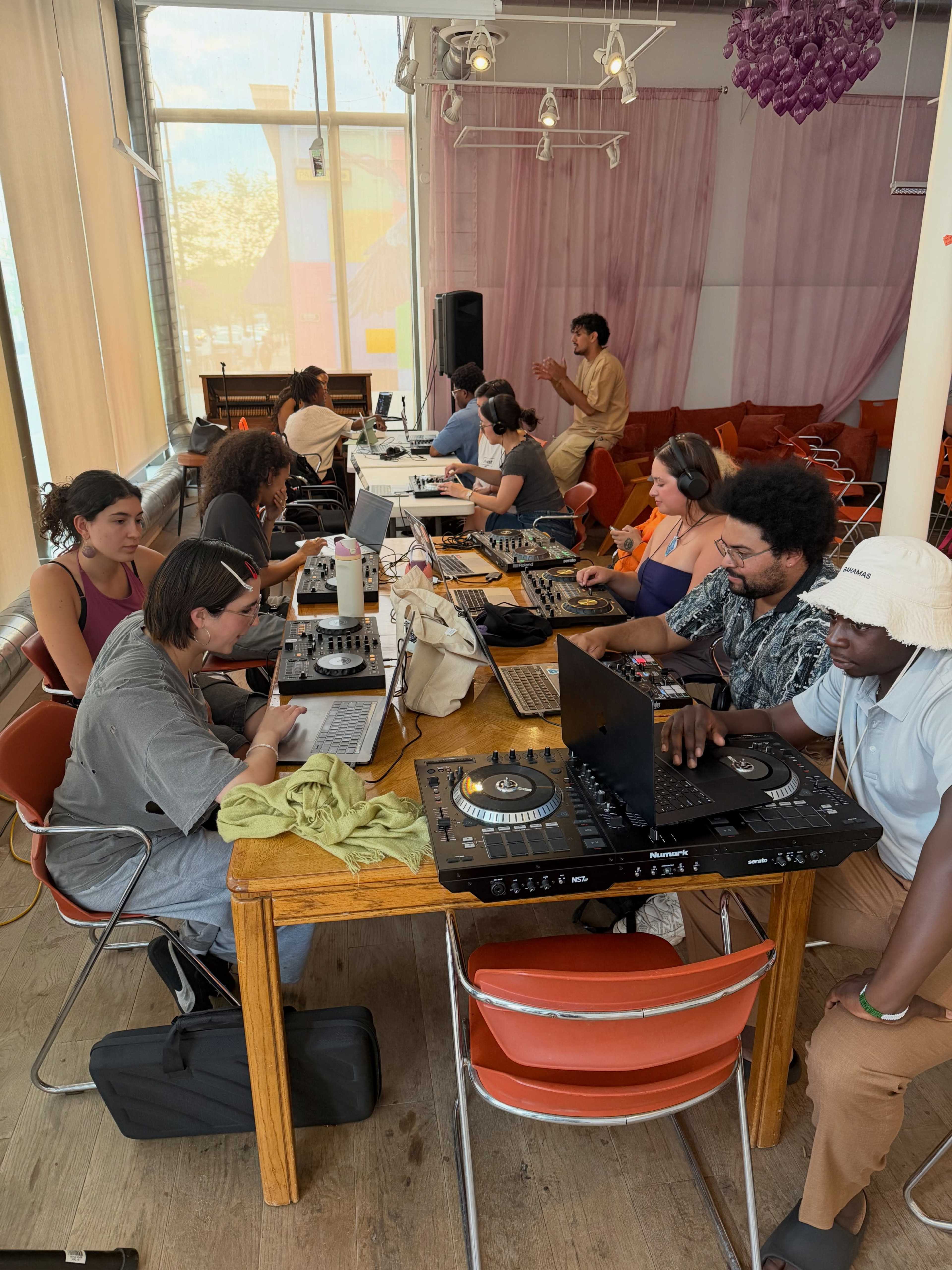 A group of people is seated around tables with electronic music equipment, engaged in a collaborative workshop or session in a well-lit indoor space.