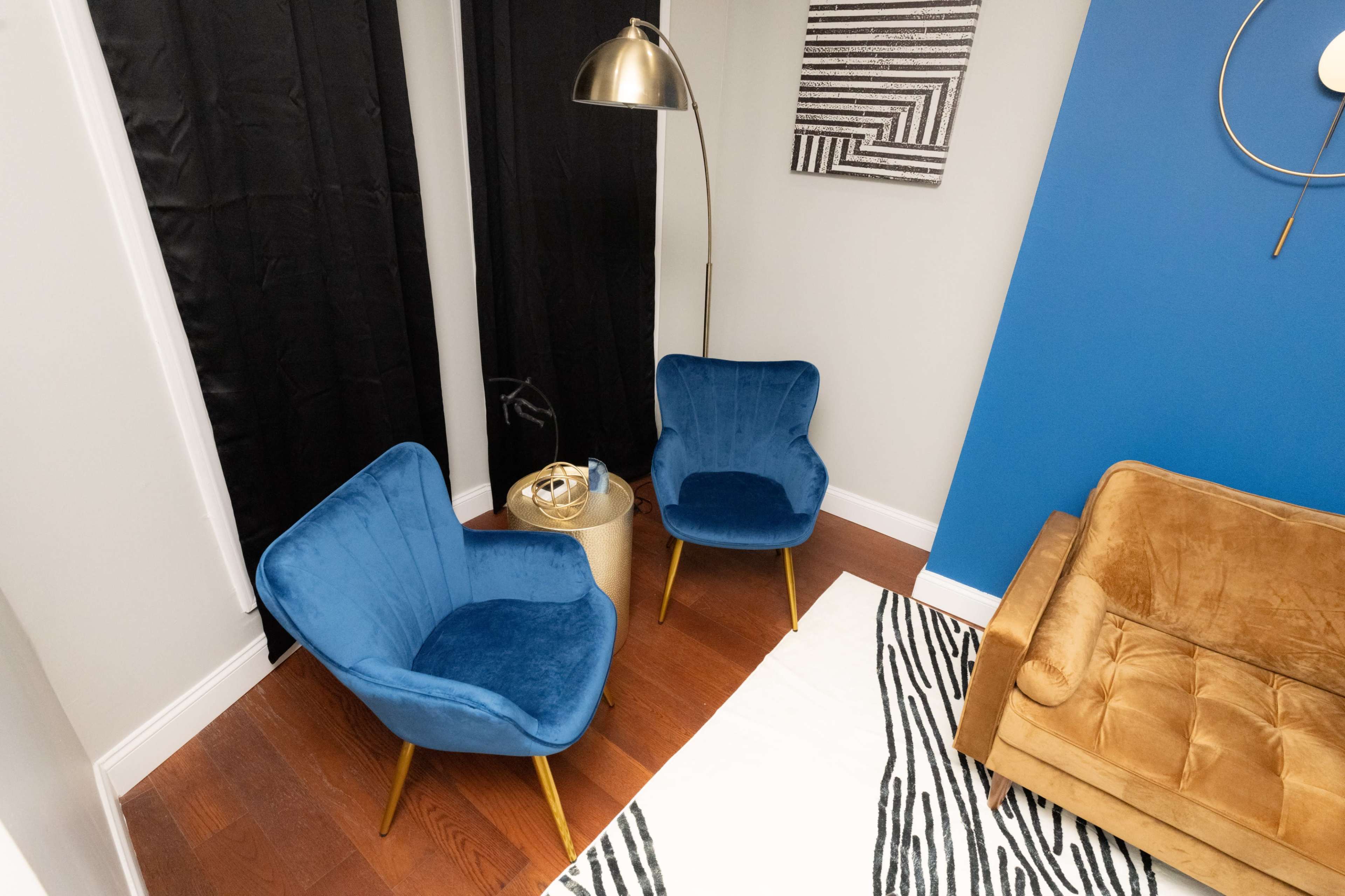 The image shows a modern living space with two blue chairs, a gold floor lamp, and a tan couch, all arranged on a striped rug.