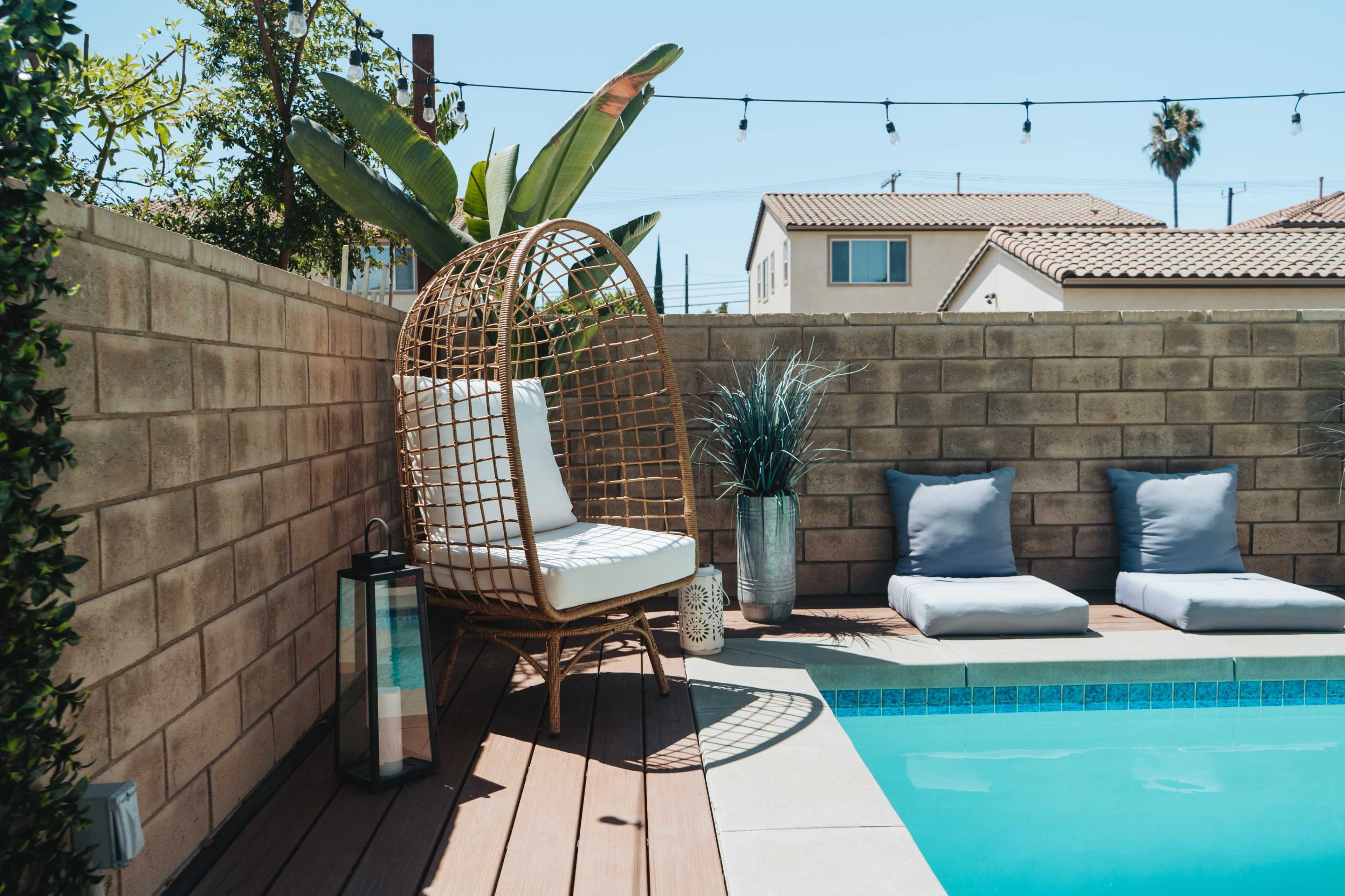 A wicker chair sits next to a swimming pool surrounded by potted plants and cushioned lounge chairs.