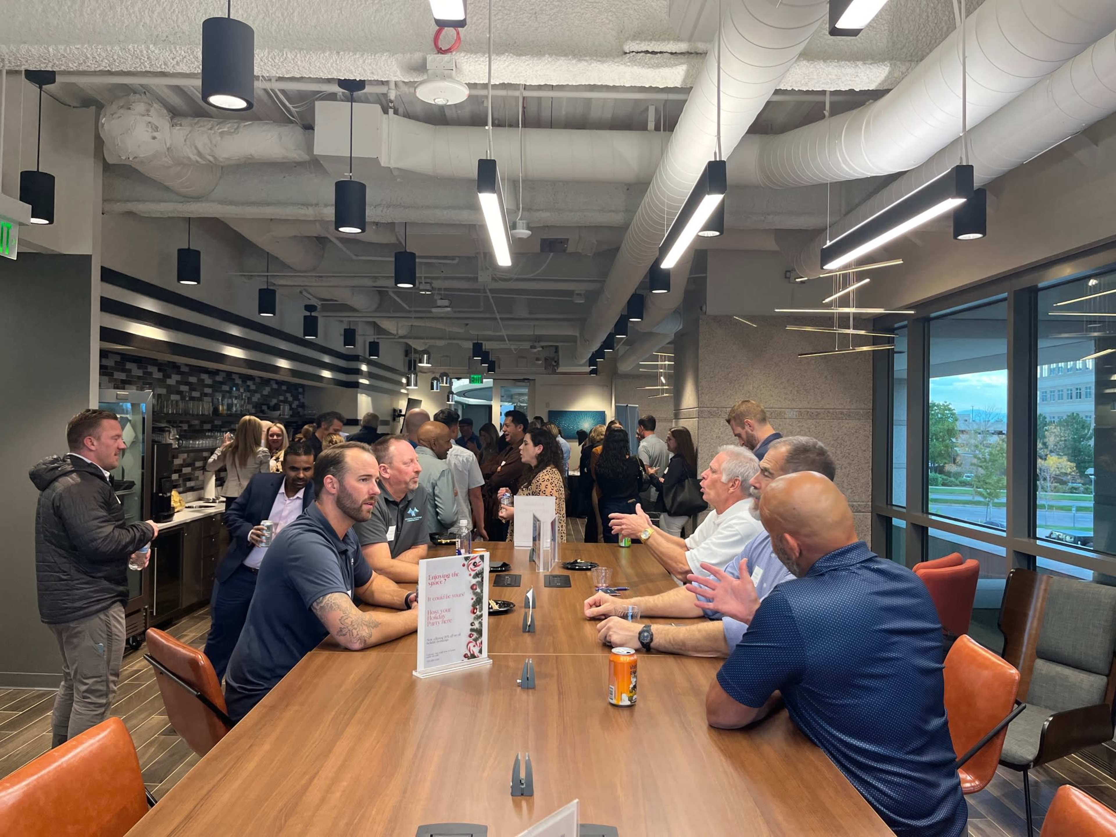 A crowded communal space features people engaged in conversation around a long table, with some individuals at a counter in the background.