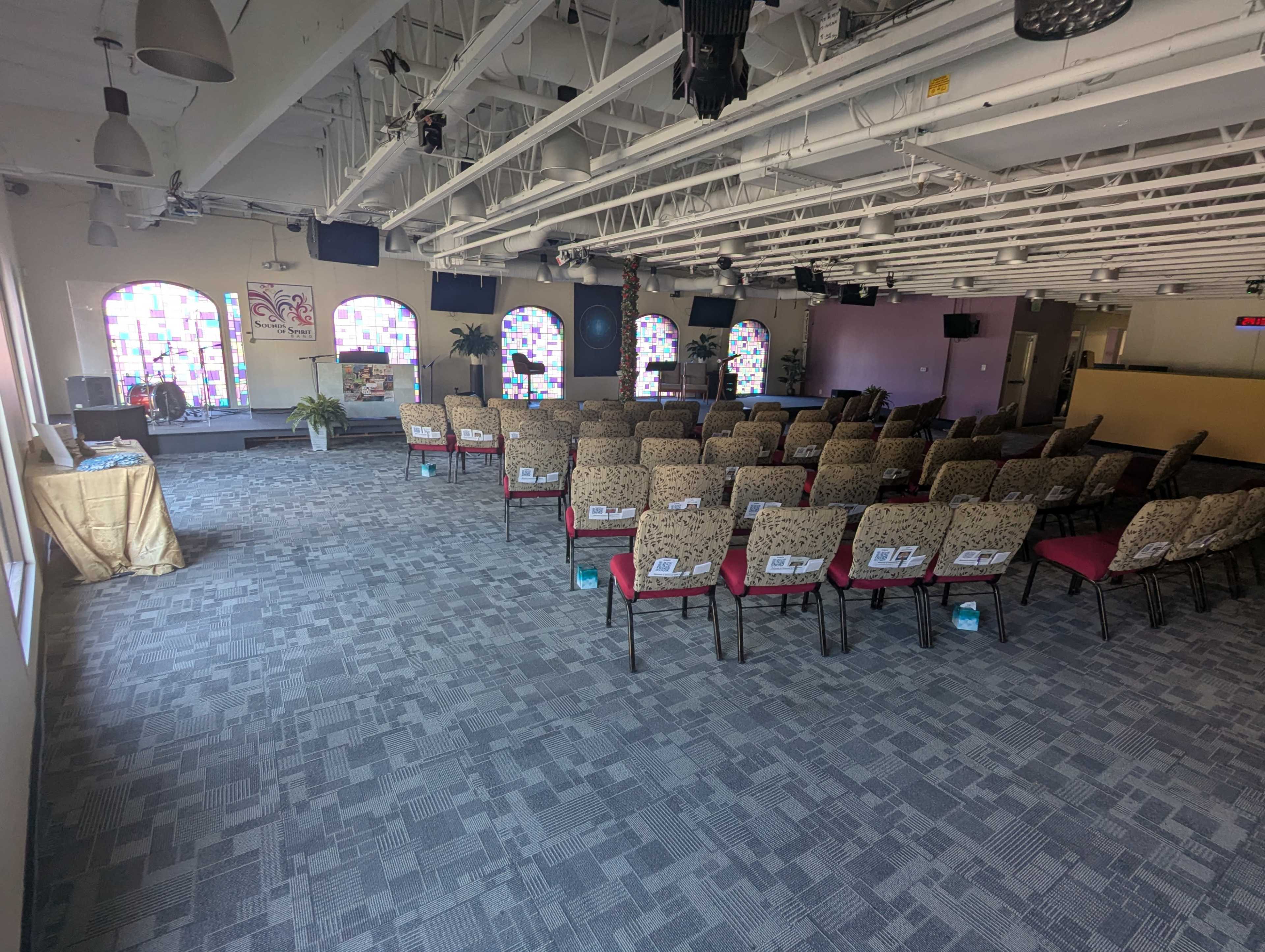 The image shows a spacious room set up with rows of chairs facing a stage, with large windows featuring colorful panes.