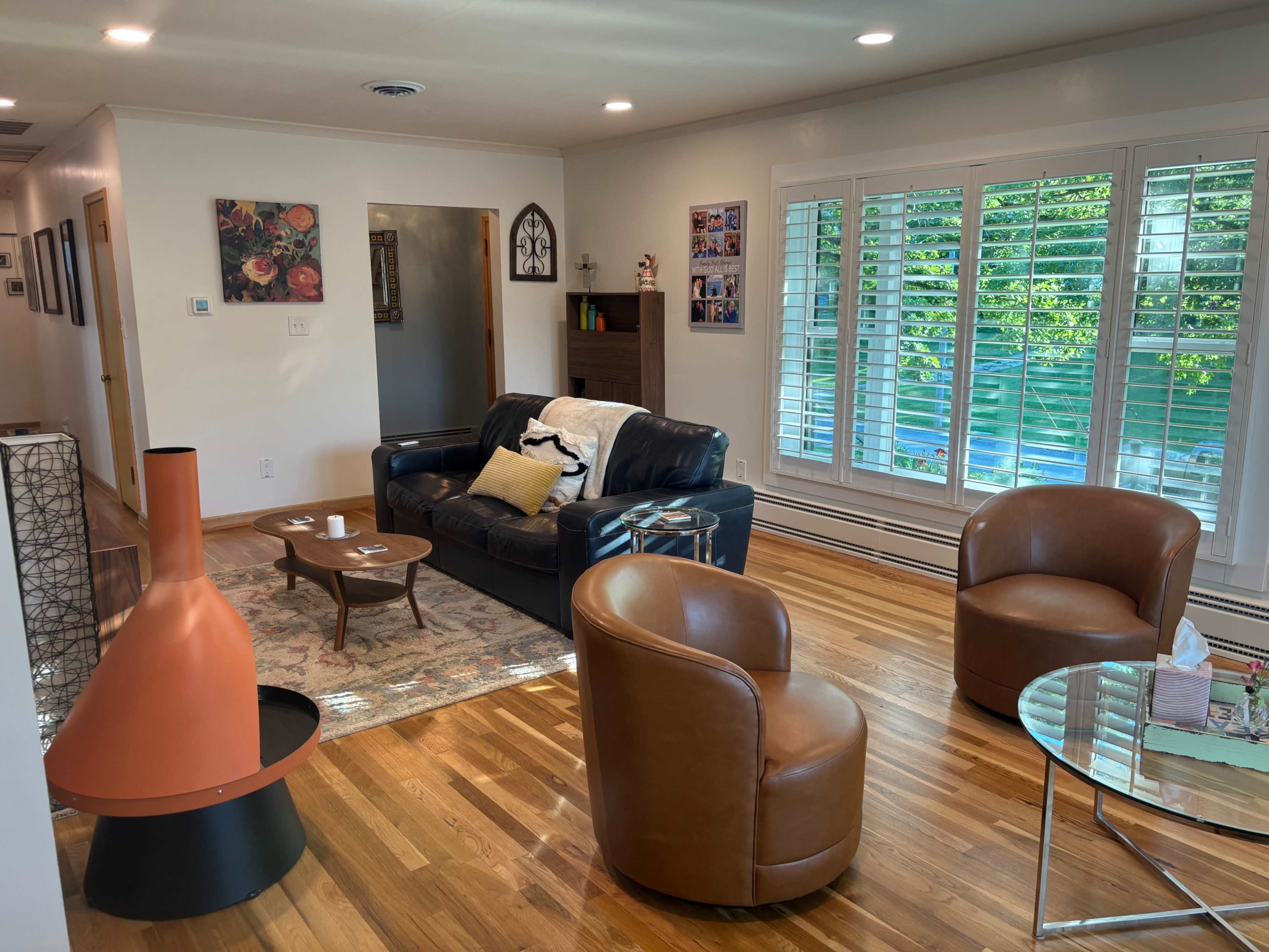 The living room features a black leather sofa, two round brown chairs, a glass coffee table, and a wooden sideboard, with large windows covered by white shutters allowing natural light to enter.