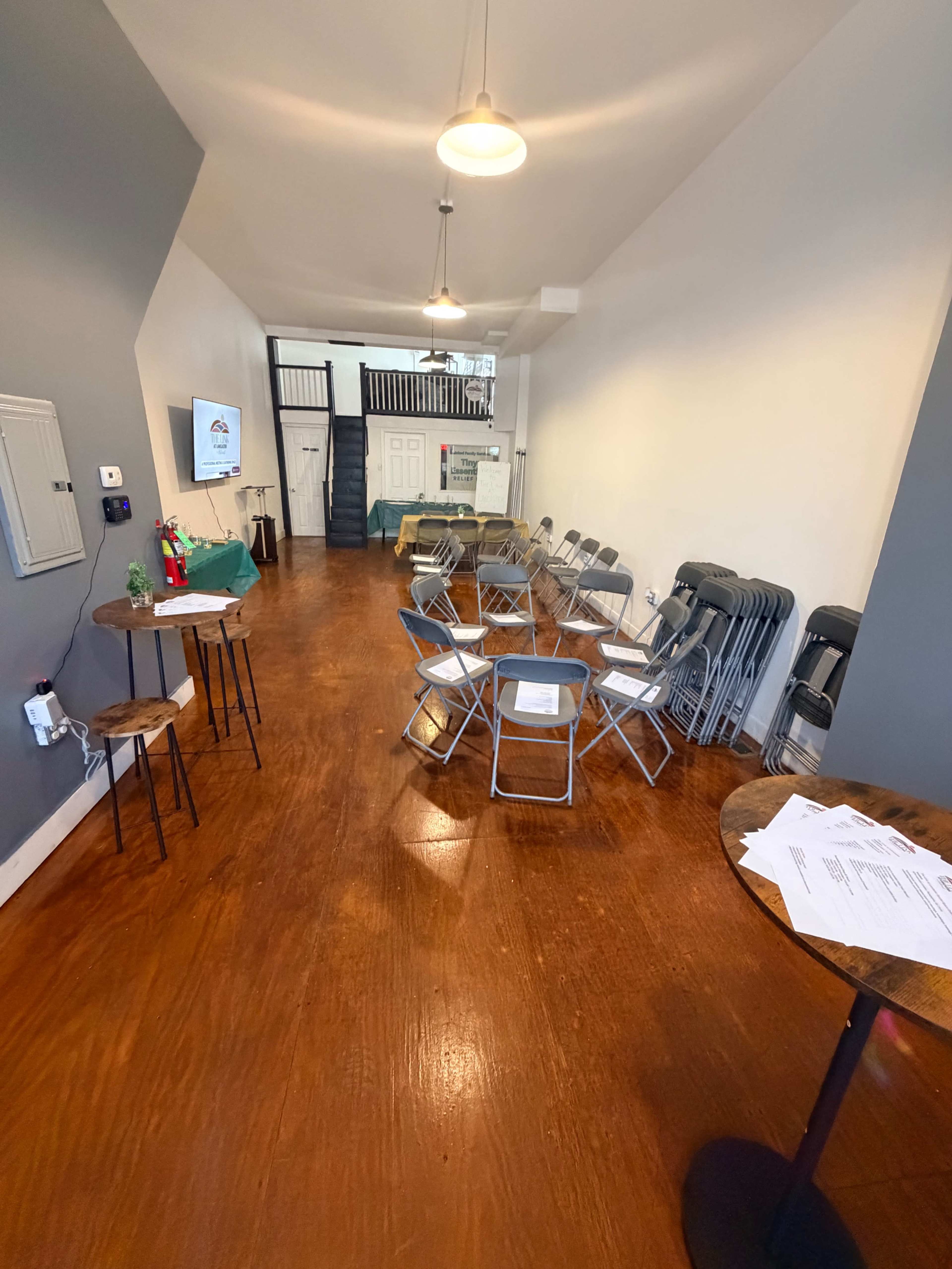 The image shows a spacious room set up for a meeting, featuring rows of folding chairs along with small tables, a television on one wall, and a staircase in the background.