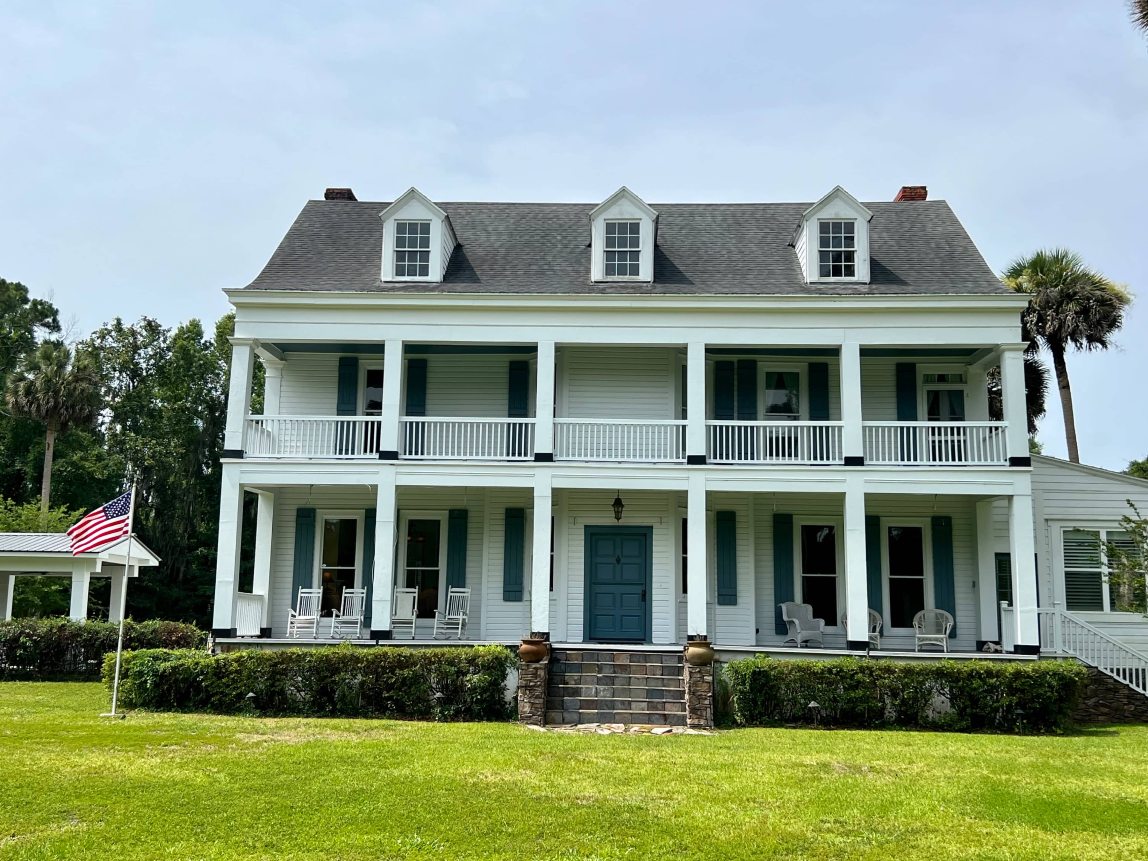A large, two-story white house with a blue front door and a porch supported by columns is set on a grassy lawn.