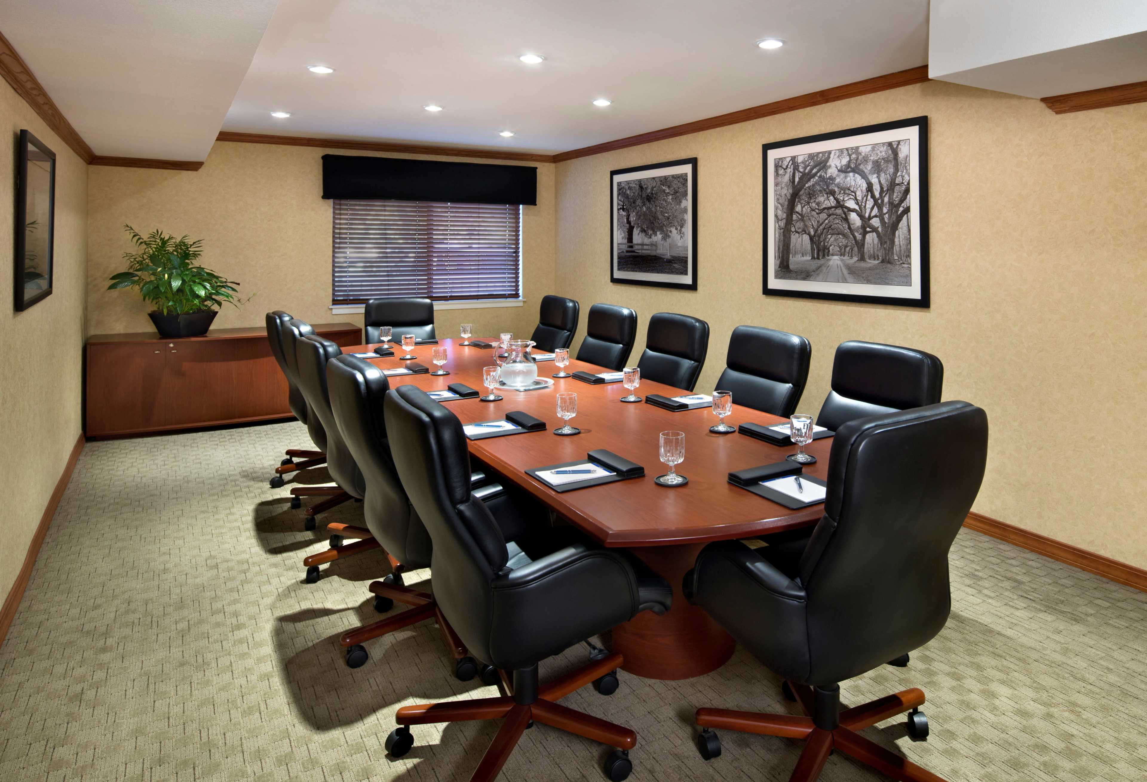 A large conference room features a long wooden table surrounded by black leather chairs, with windows covered by blinds and framed black-and-white photographs on the walls.