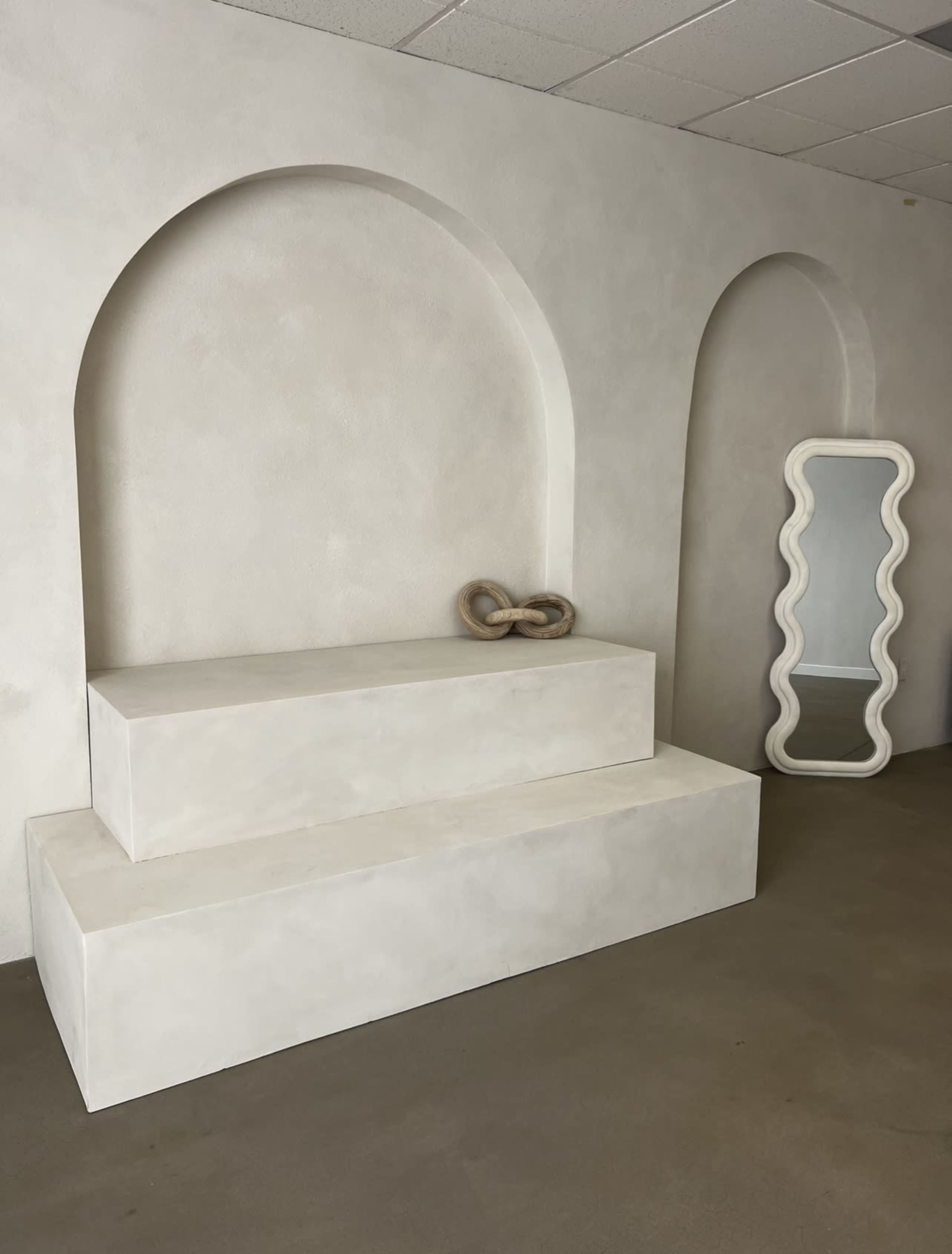 A minimalist room features a white, tiered platform and a wavy-edged mirror against a textured wall with arched niches.