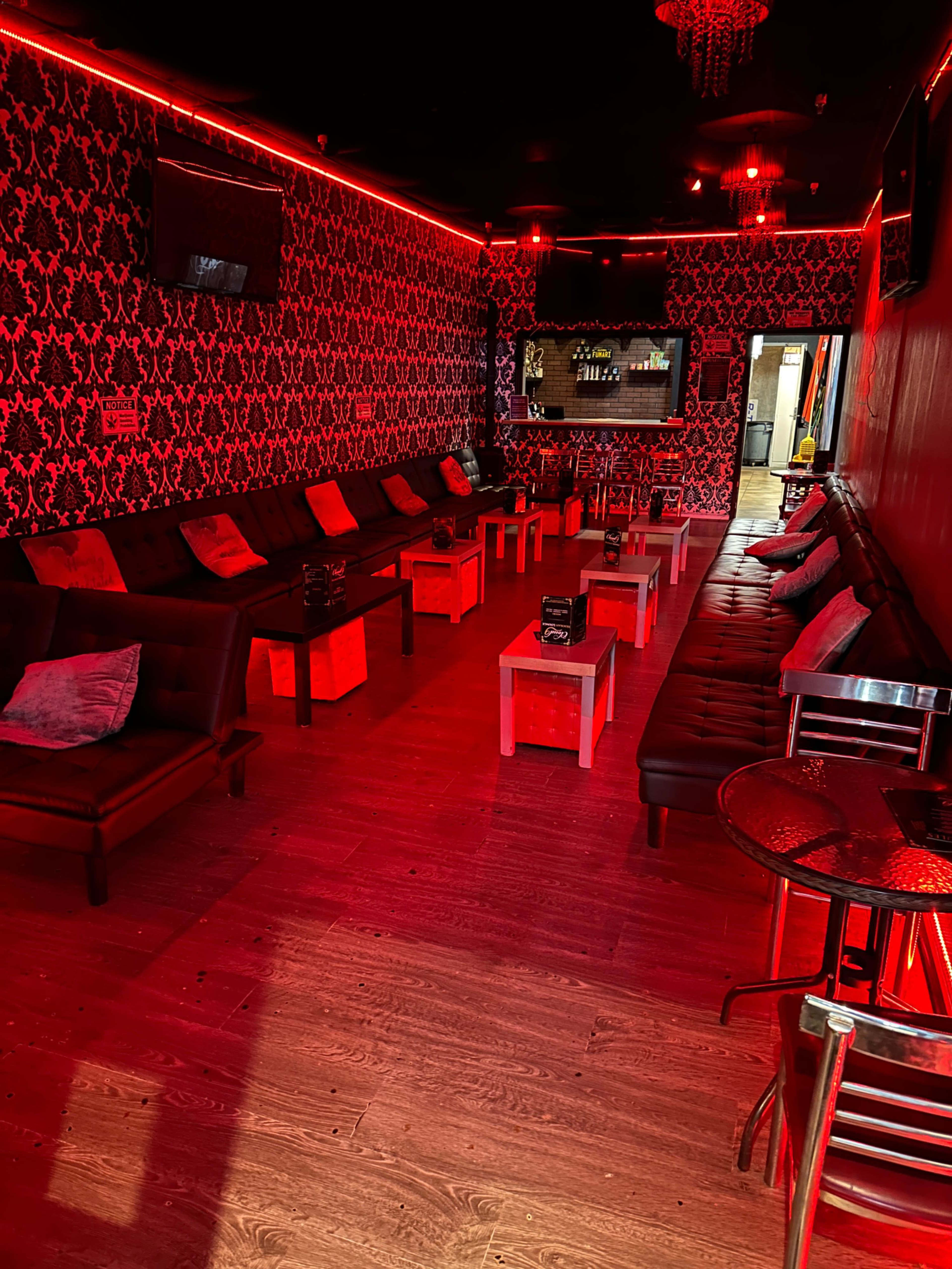 A dimly lit lounge with red lighting, featuring black couches arranged around small tables on a wooden floor.
