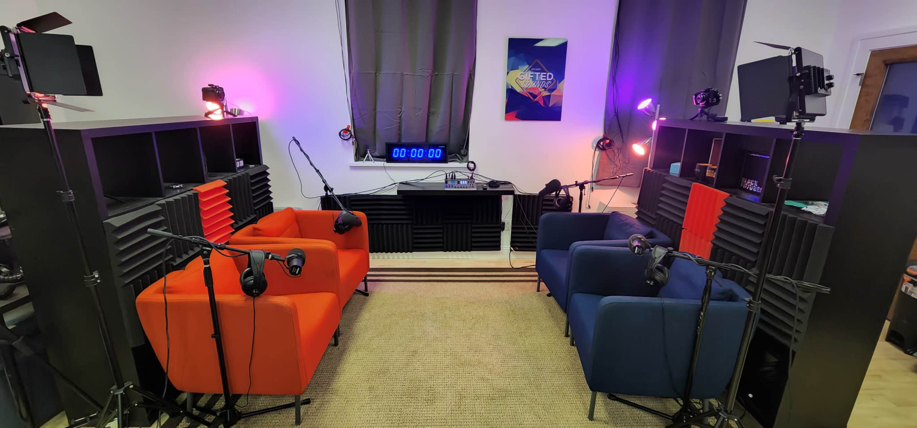 A recording studio features two color-coded seating areas, surrounded by microphones and lighting equipment, with a large digital clock on the wall.
