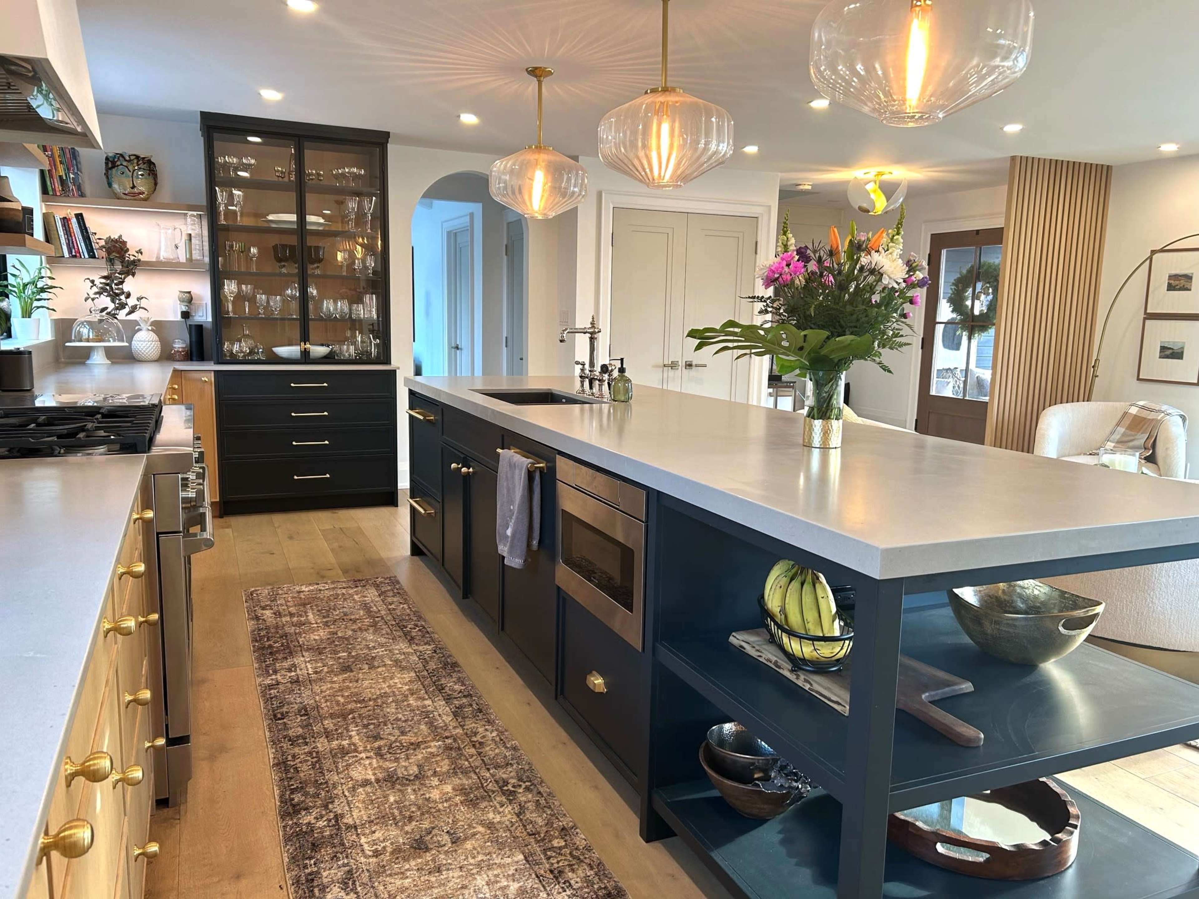 A modern kitchen features a central island with a countertop, a gas stove, dark cabinetry, glass-front shelves displaying dishware, and decorative plants.