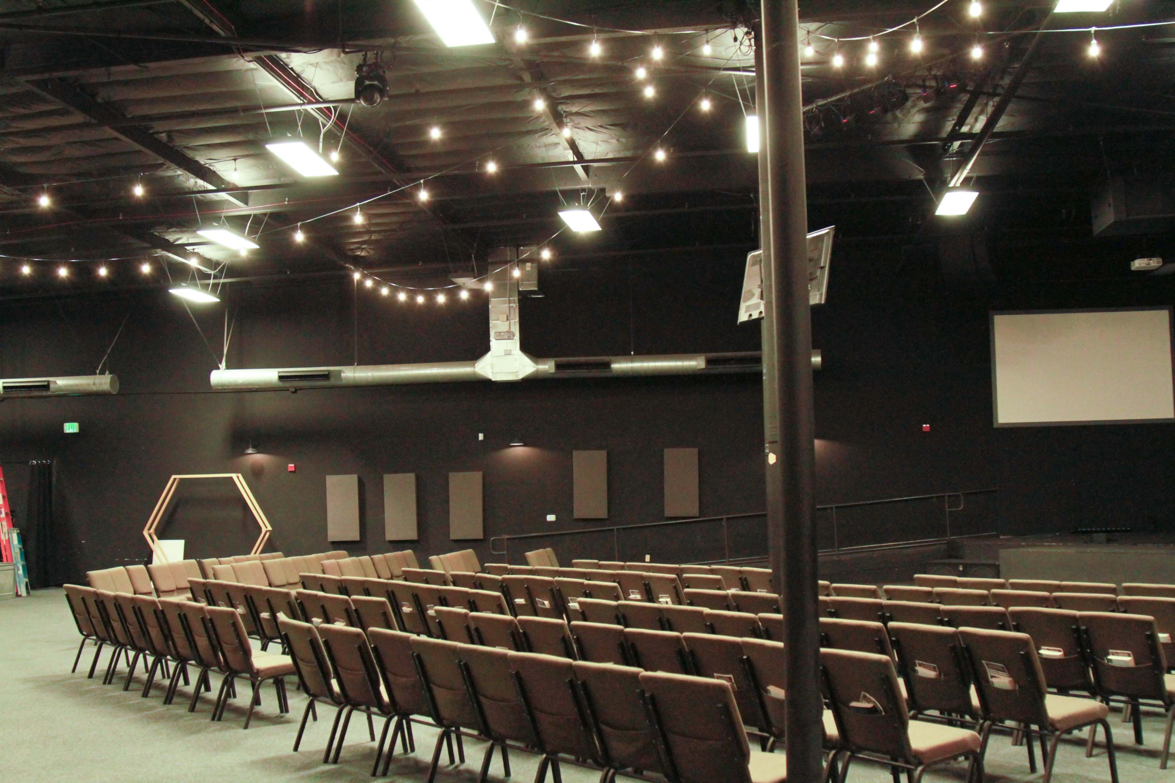 The image shows an interior space set up with rows of brown chairs facing a staged area, illuminated by string lights overhead.