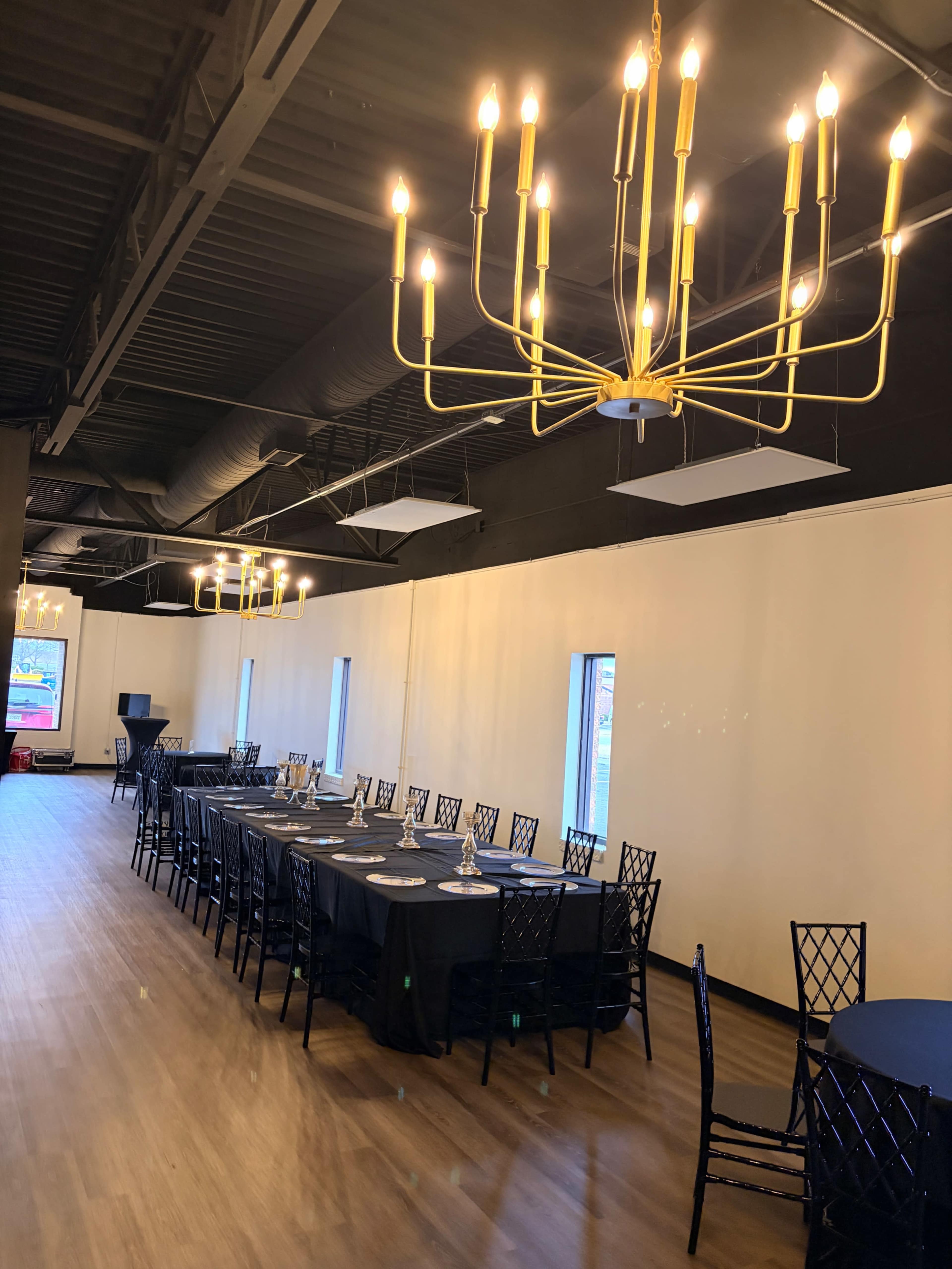 The image shows a spacious event hall with a long table set for a gathering, surrounded by black chairs and illuminated by a golden chandelier.