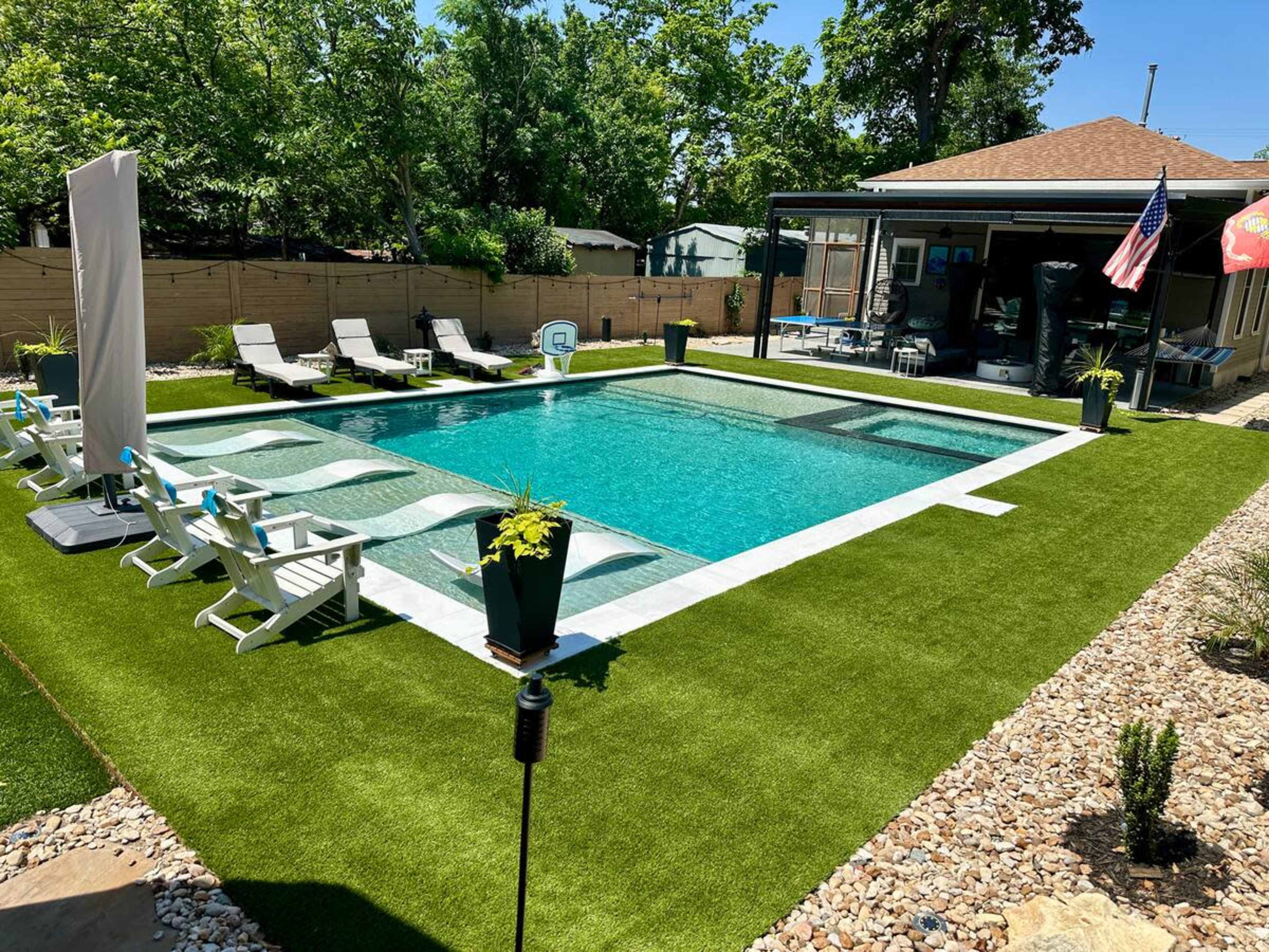 The image shows a rectangular swimming pool surrounded by green artificial grass, lounge chairs, potted plants, and a covered patio area.